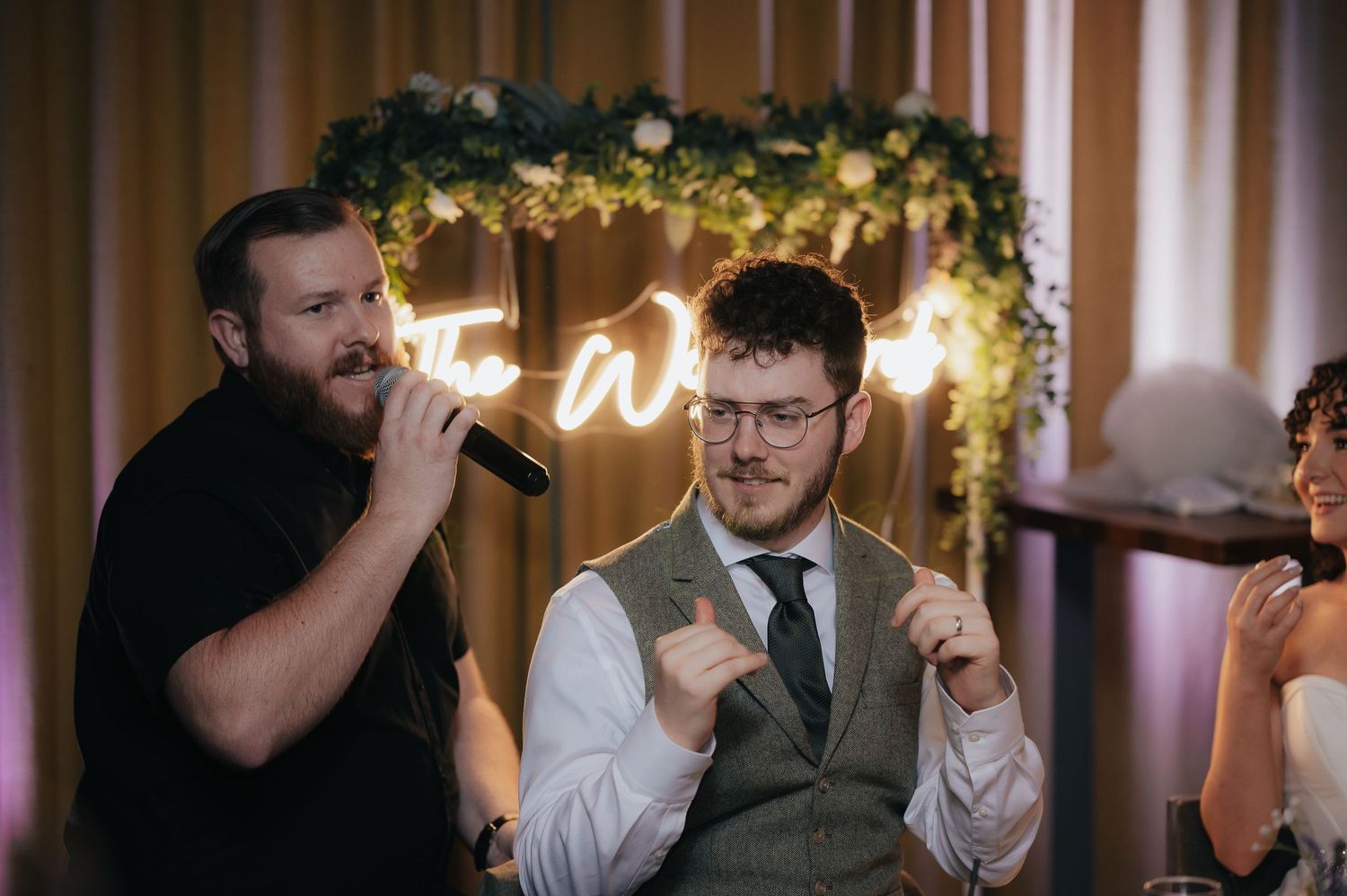 Wedding reception guests enjoy drinks under decorative lights and greenery at an indoor venue.