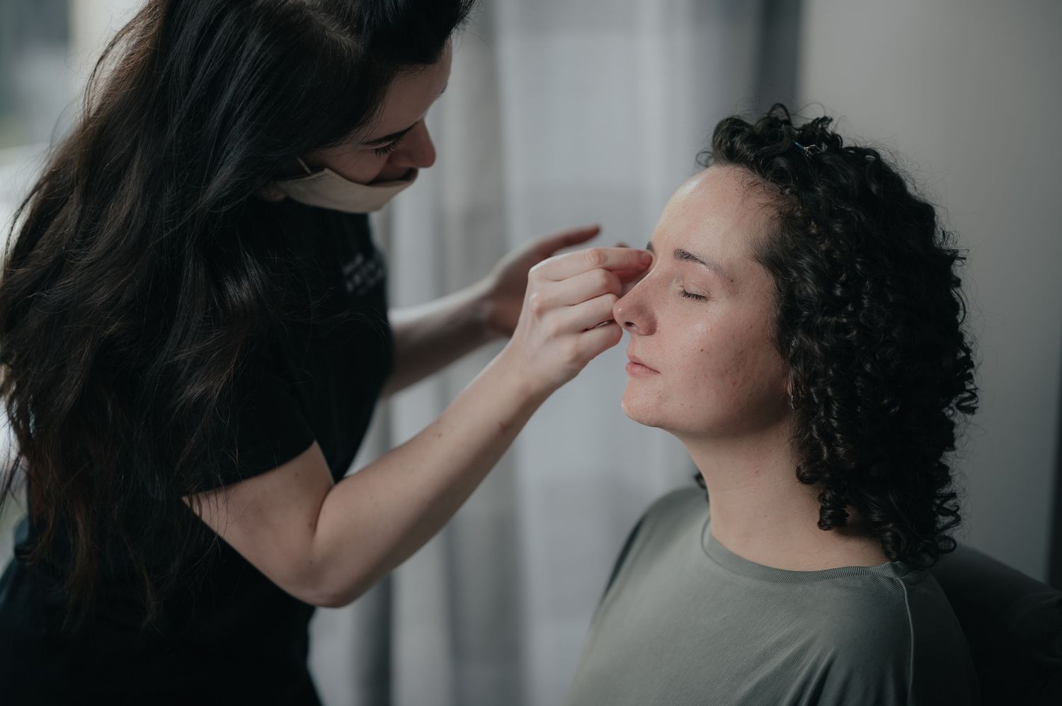 Makeup artist applying cosmetics to a client with curly hair in a neutral setting.