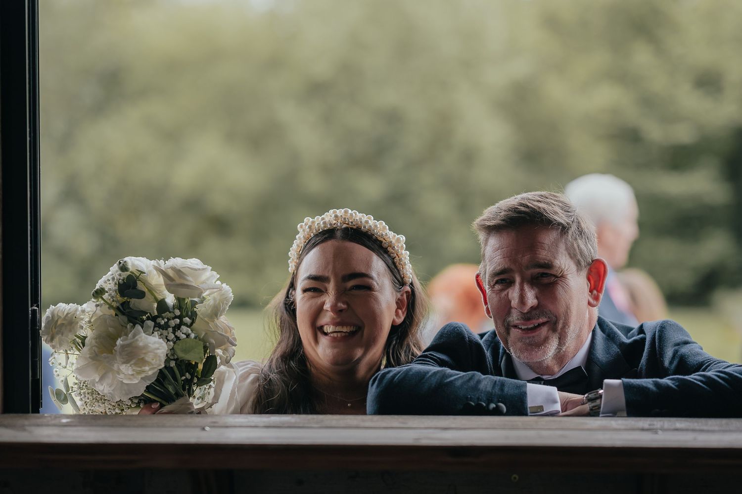 A joyful wedding scene captured through a window with a white bouquet in the foreground.