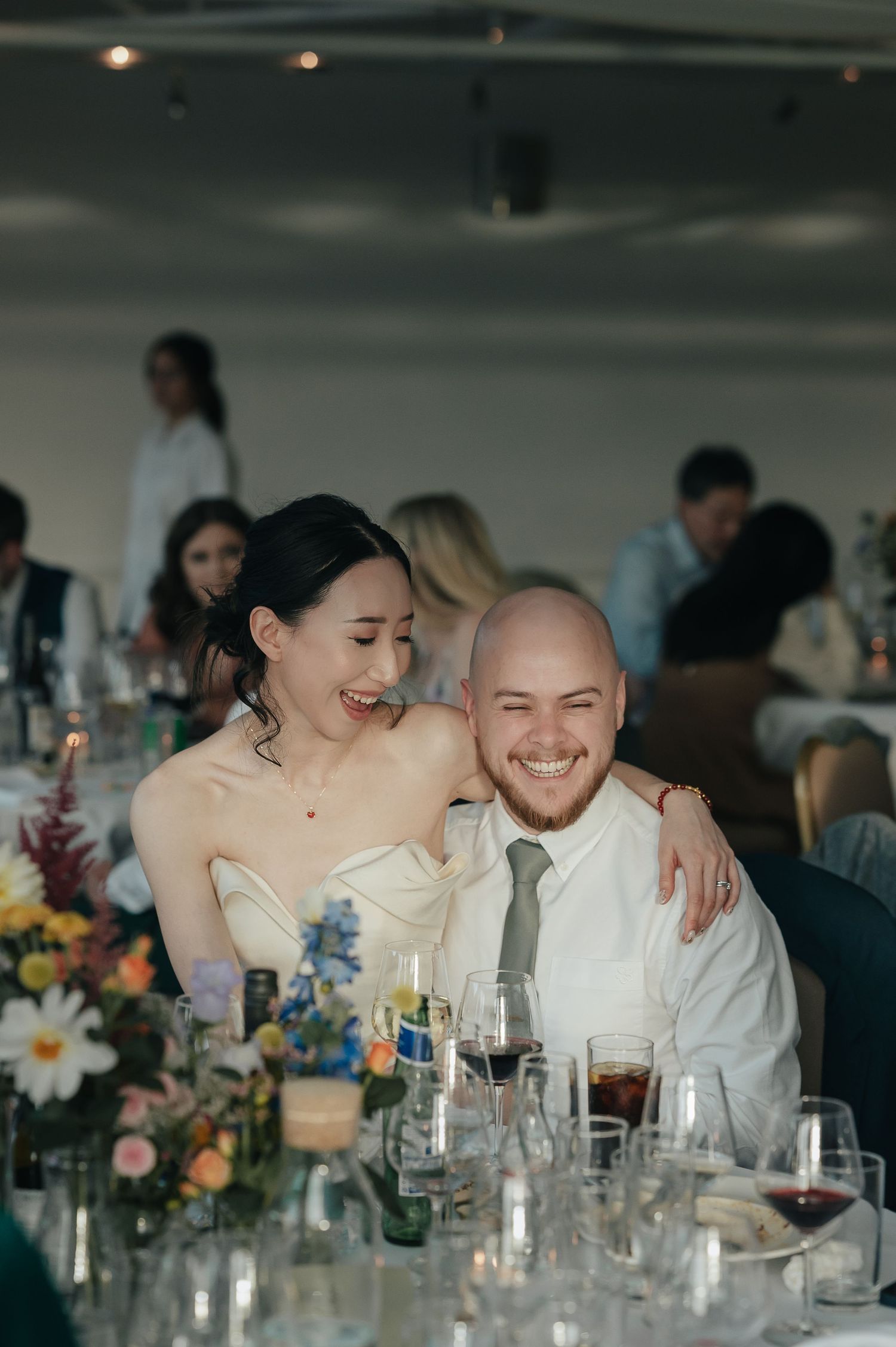 Newlyweds share joyful moment at reception dinner table surrounded by floral arrangements.
