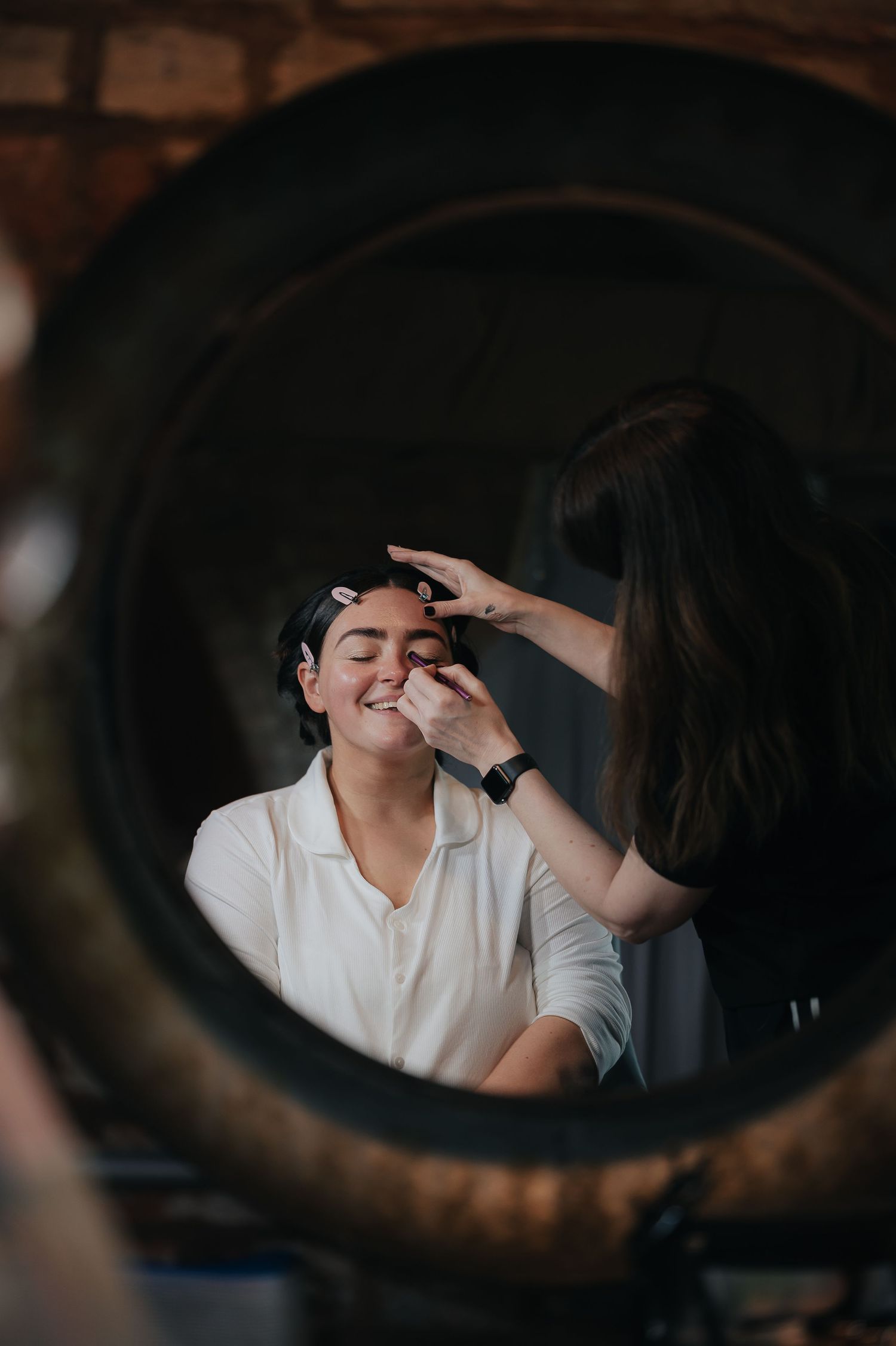 A reflection in a round mirror shows someone getting their makeup done.