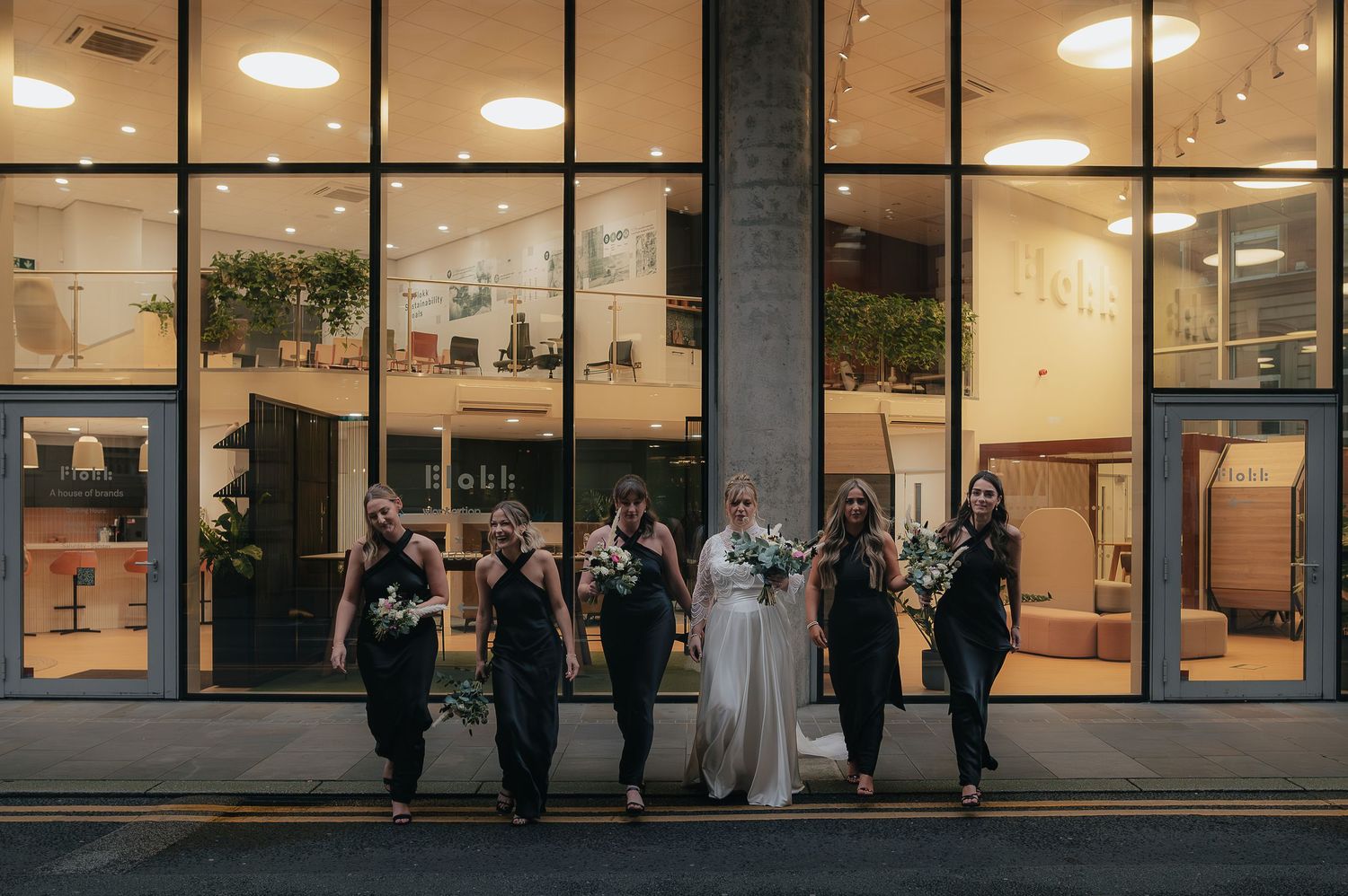A wedding party poses outside illuminated storefront windows at night with warm interior lighting glowing behind them.