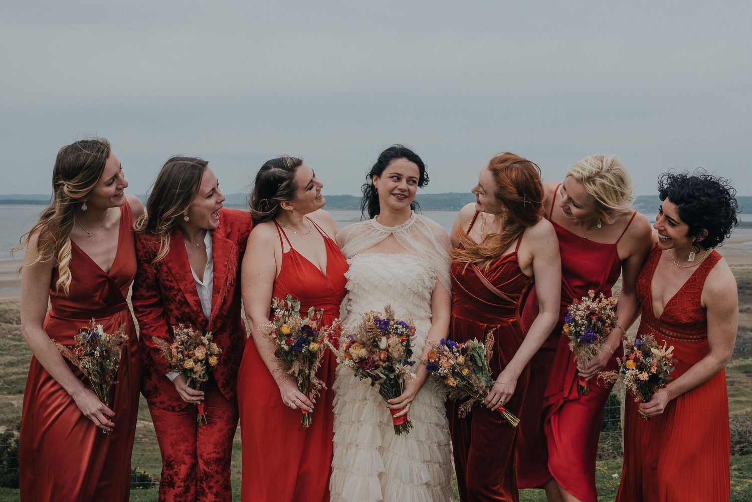 Bridal party in red dresses poses with bouquets against coastal backdrop.