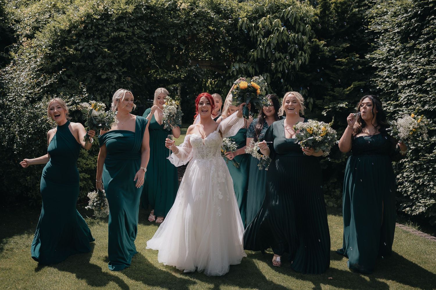 Bridal party in emerald green dresses posing with bride in garden setting.