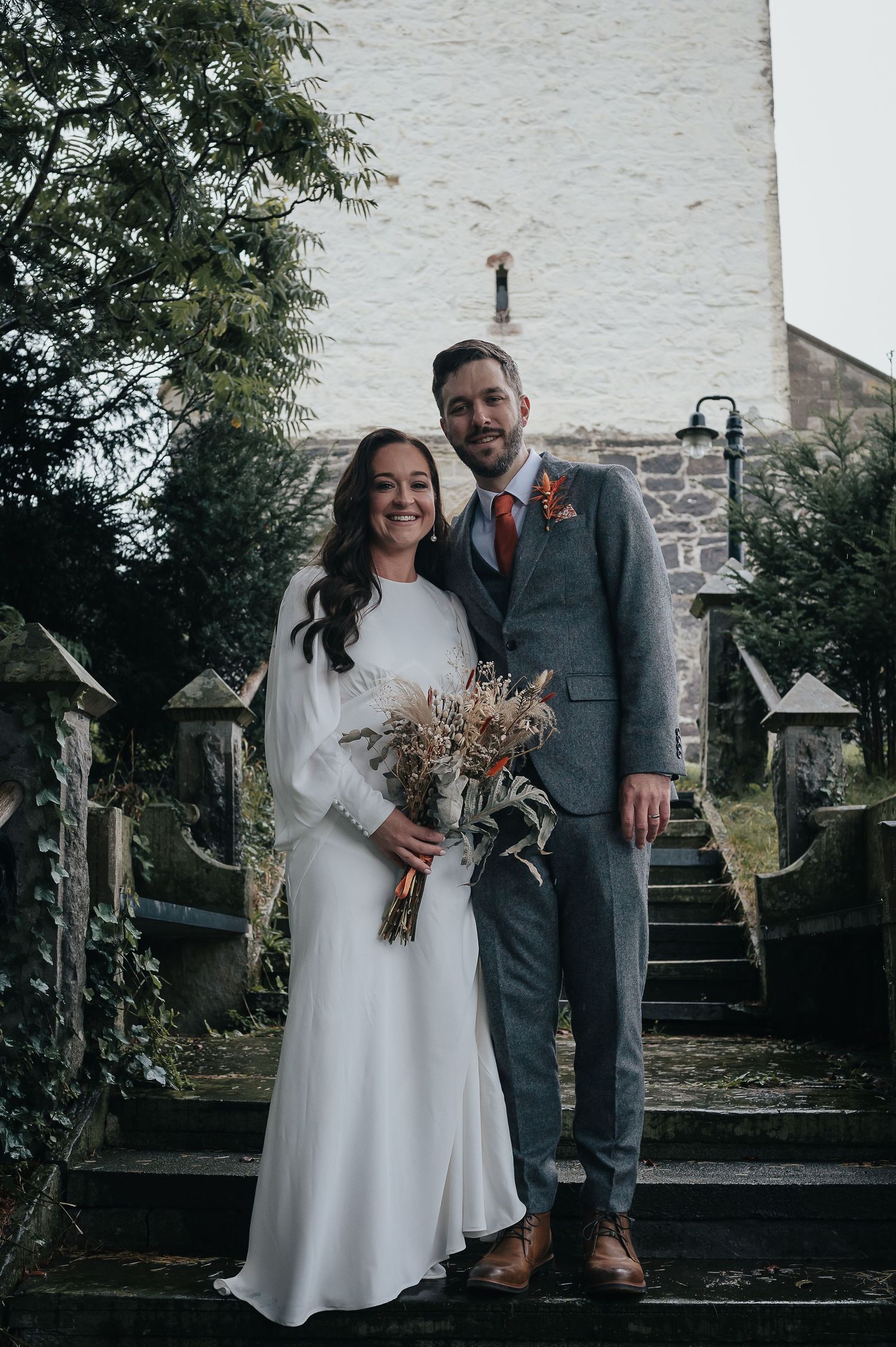 A couple poses on stone steps with dried flower bouquet at their wedding.