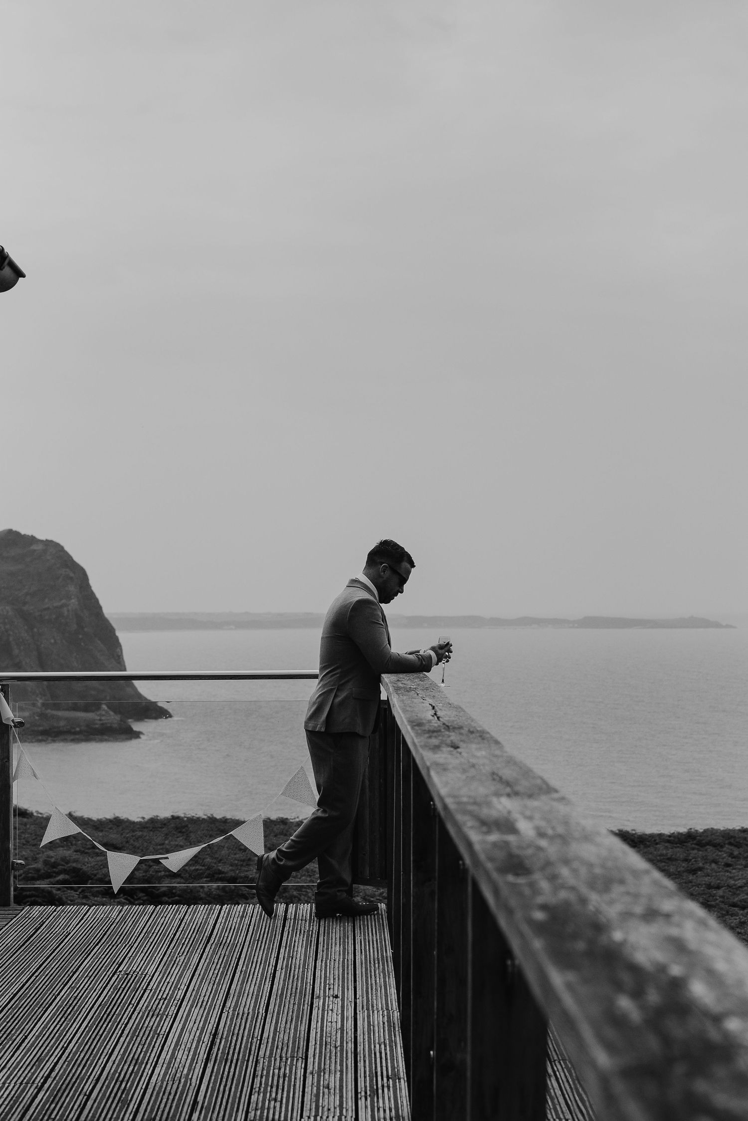 Person in formal attire standing on a deck overlooking coastal cliffs in black and white.
