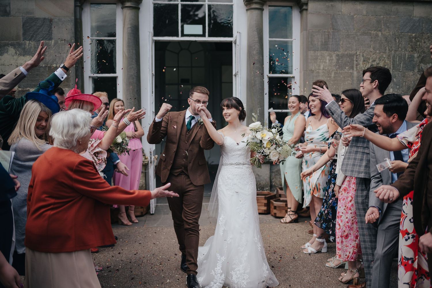 Wedding guests shower the newlyweds with confetti outside a historic stone building during an energetic celebration.