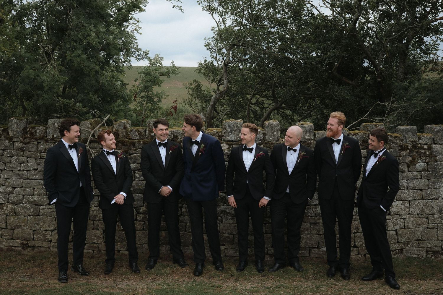 Wedding party members in formal attire lined up against a stone wall backdrop.