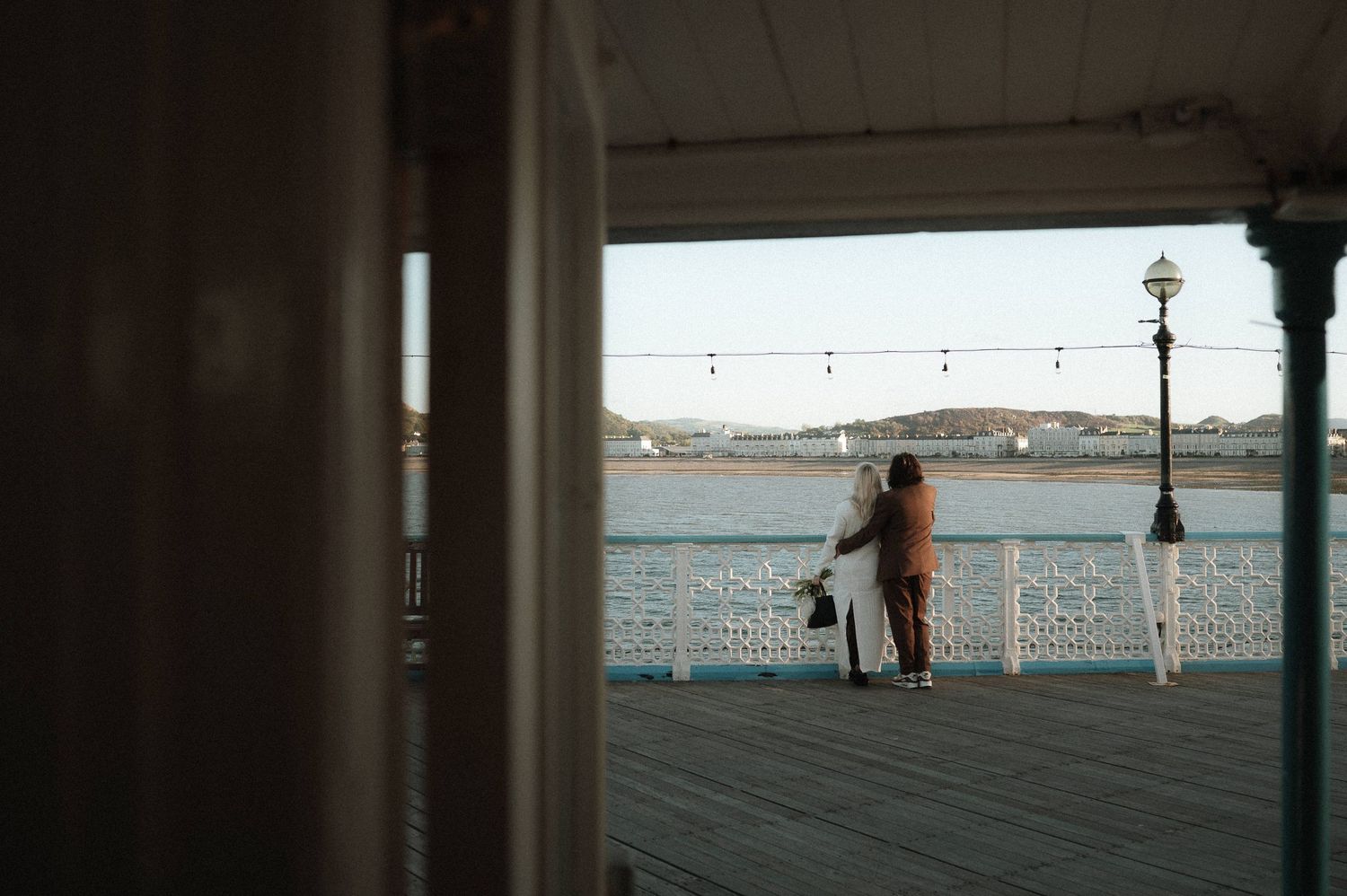 A sequence of silhouetted figures walking along a waterfront boardwalk against a bright horizon.