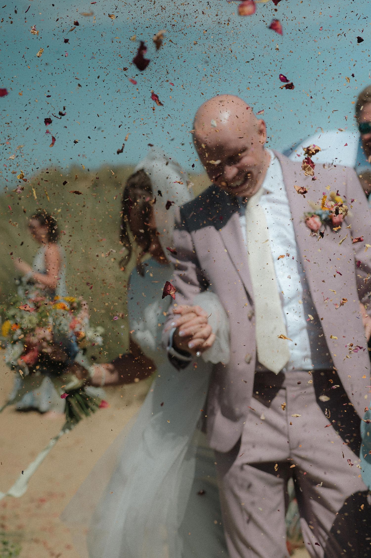 A couple celebrates their wedding moment as colorful confetti rains down around them against a turquoise background.