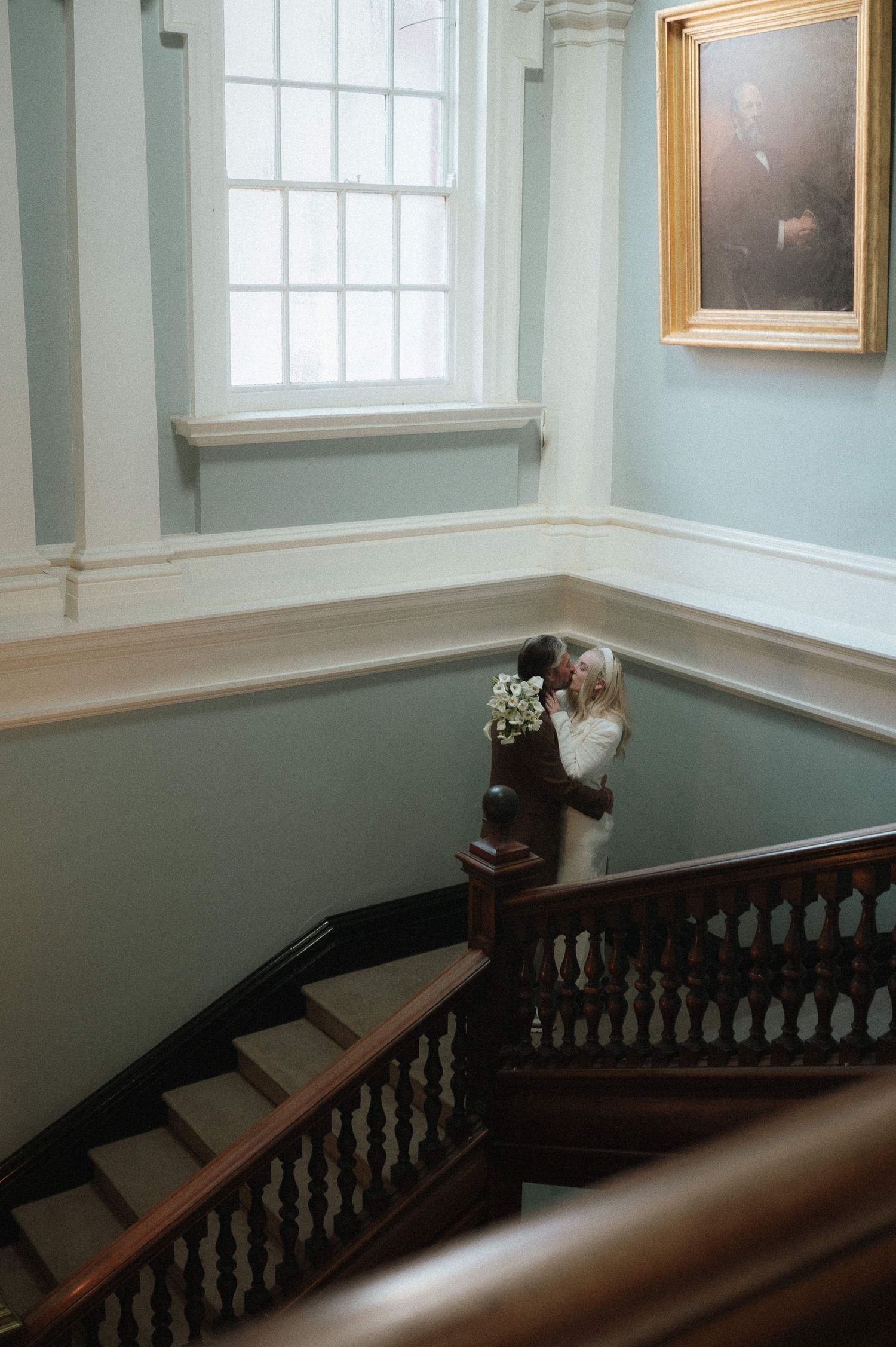 Historic staircase with dark wooden banister and mint green walls in a colonial style building with natural light from windows.