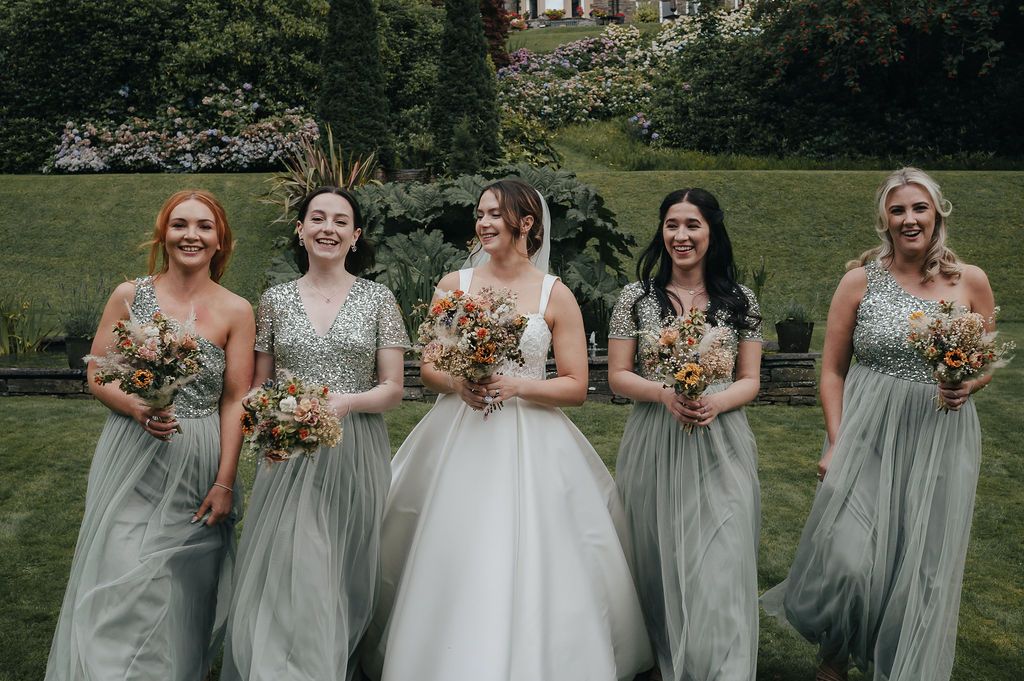 Wedding party in sage green dresses holding floral bouquets on a mansion lawn.