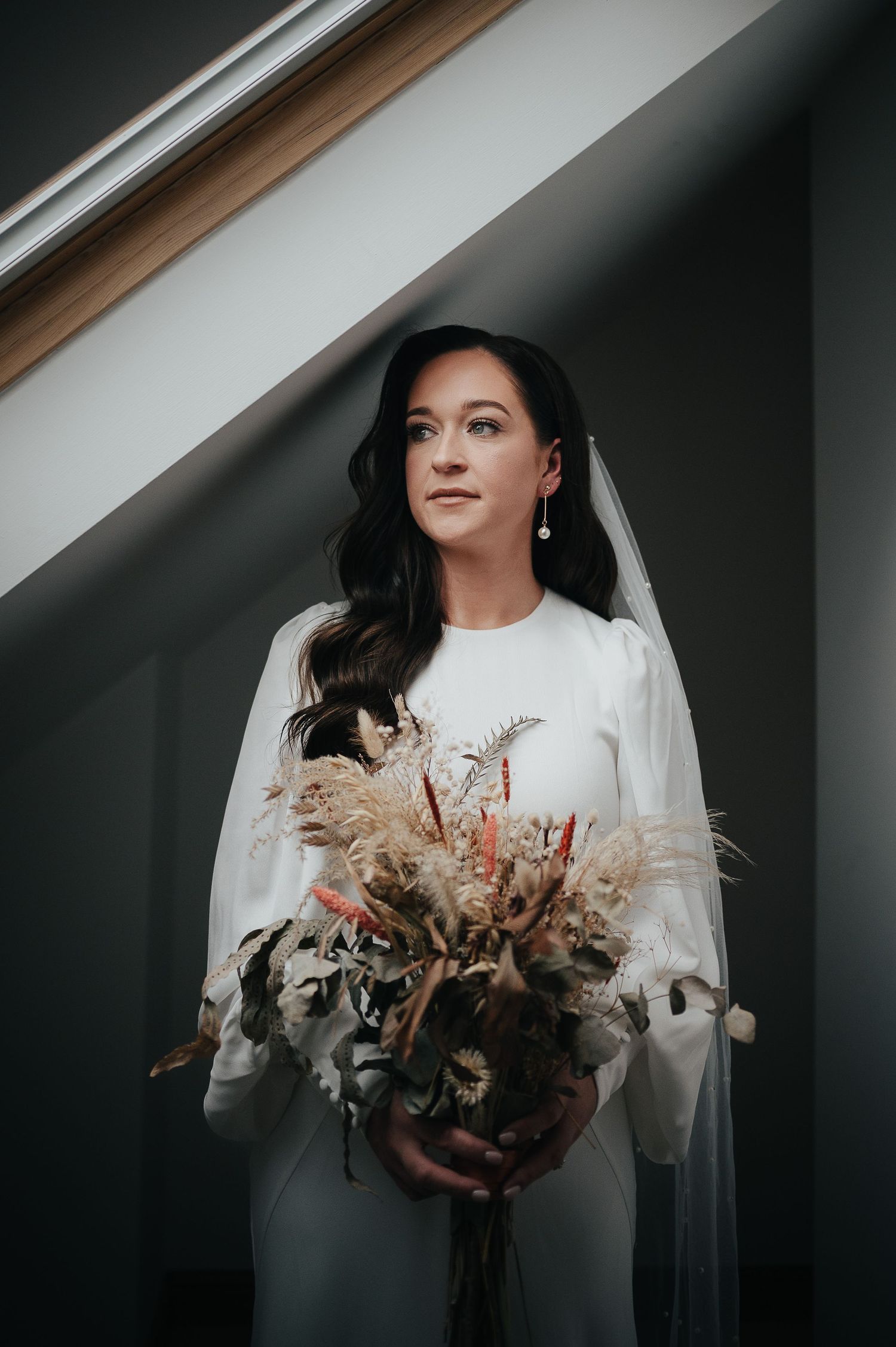A bride in a modern white dress holds a stunning dried flower bouquet in moody lighting.