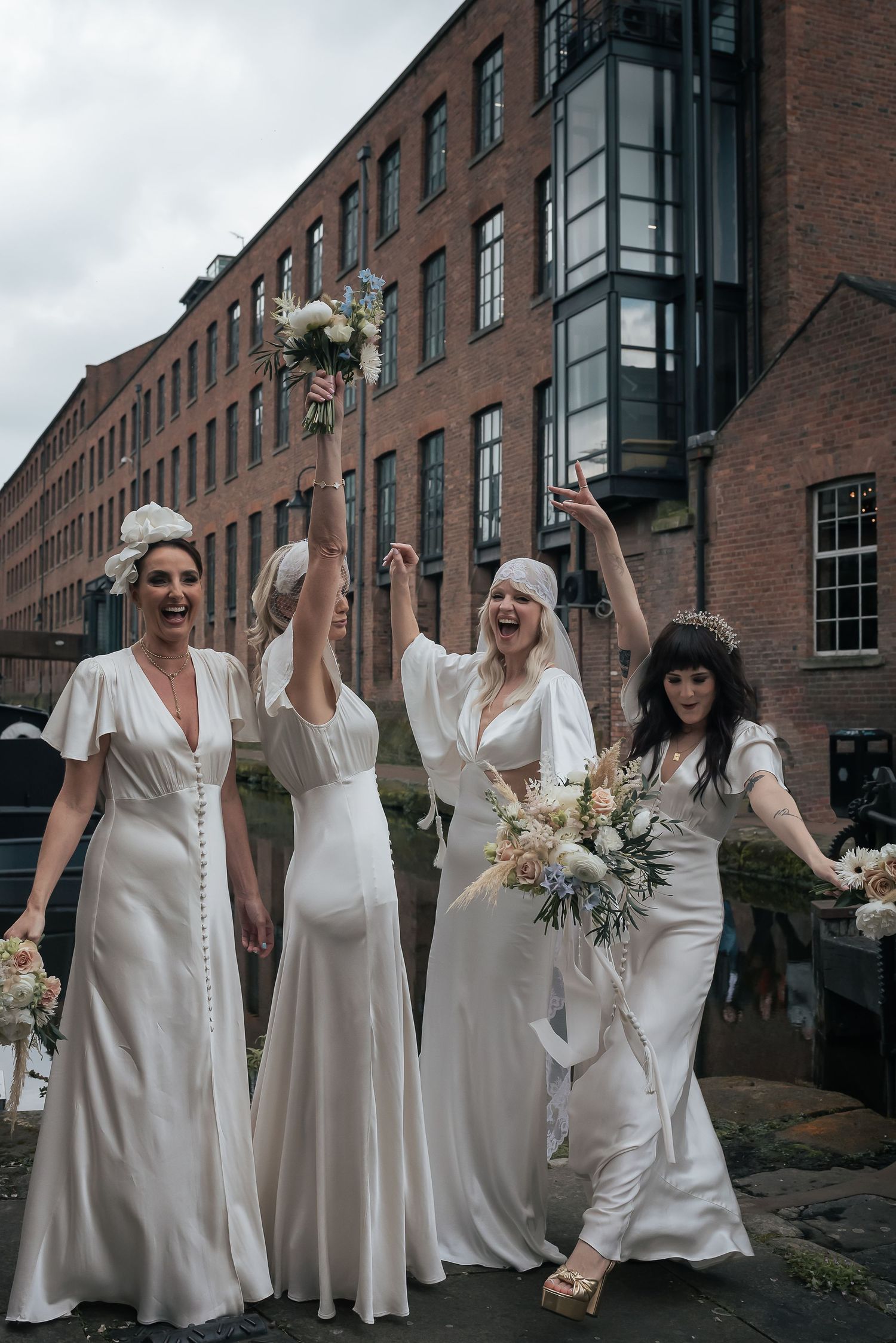 Four bridesmaids in white dresses celebrate joyfully outside brick industrial building.