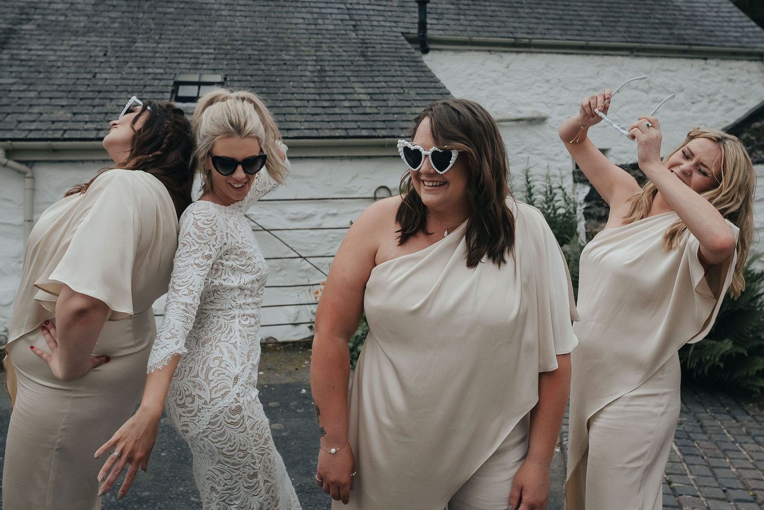 Women in light-colored dresses dancing and laughing together against a white building.
