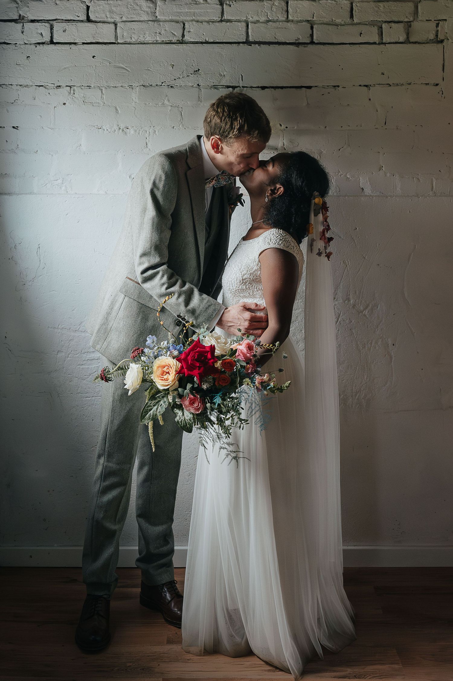A tender moment between newlyweds against a white brick wall with a colorful bouquet.