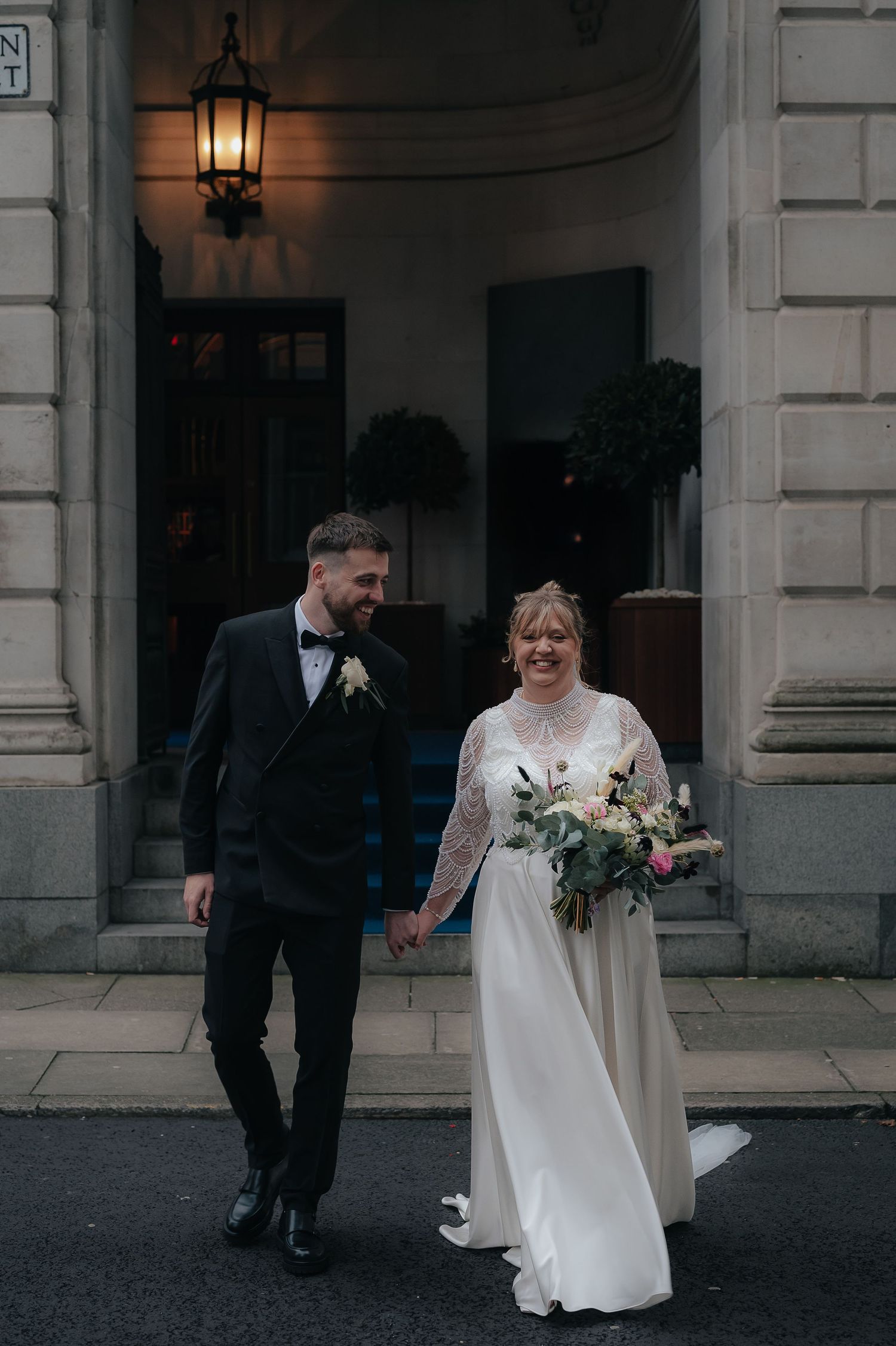 A wedding couple holding hands while walking outdoors near a traditional building entrance with a lit lantern.