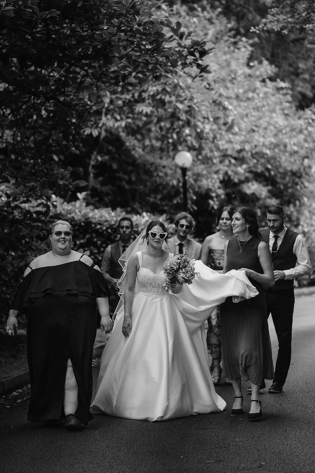 Wedding party walks together on a tree-lined path in black and white photography.