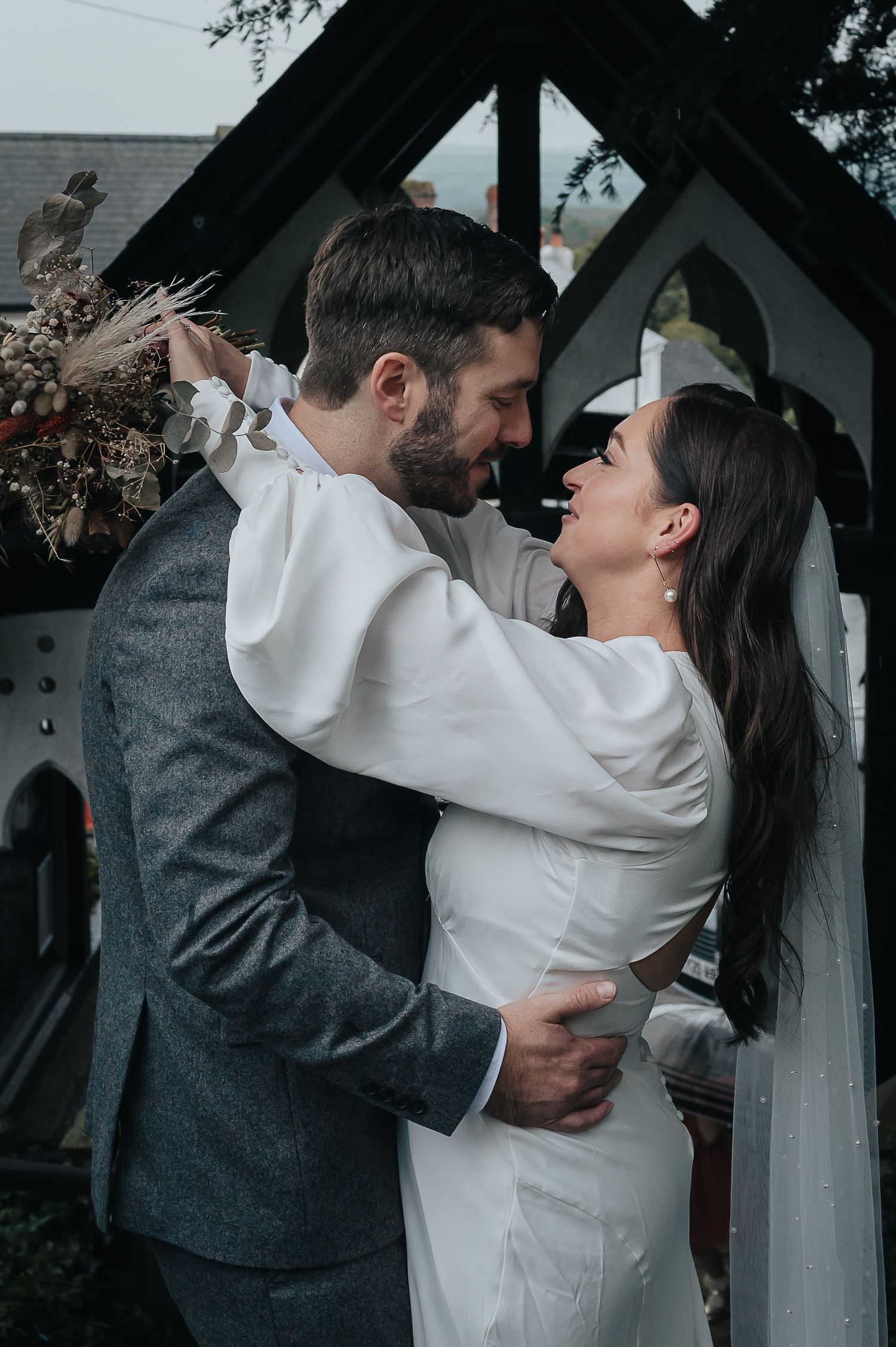 A couple in wedding attire share a romantic moment under an outdoor archway.