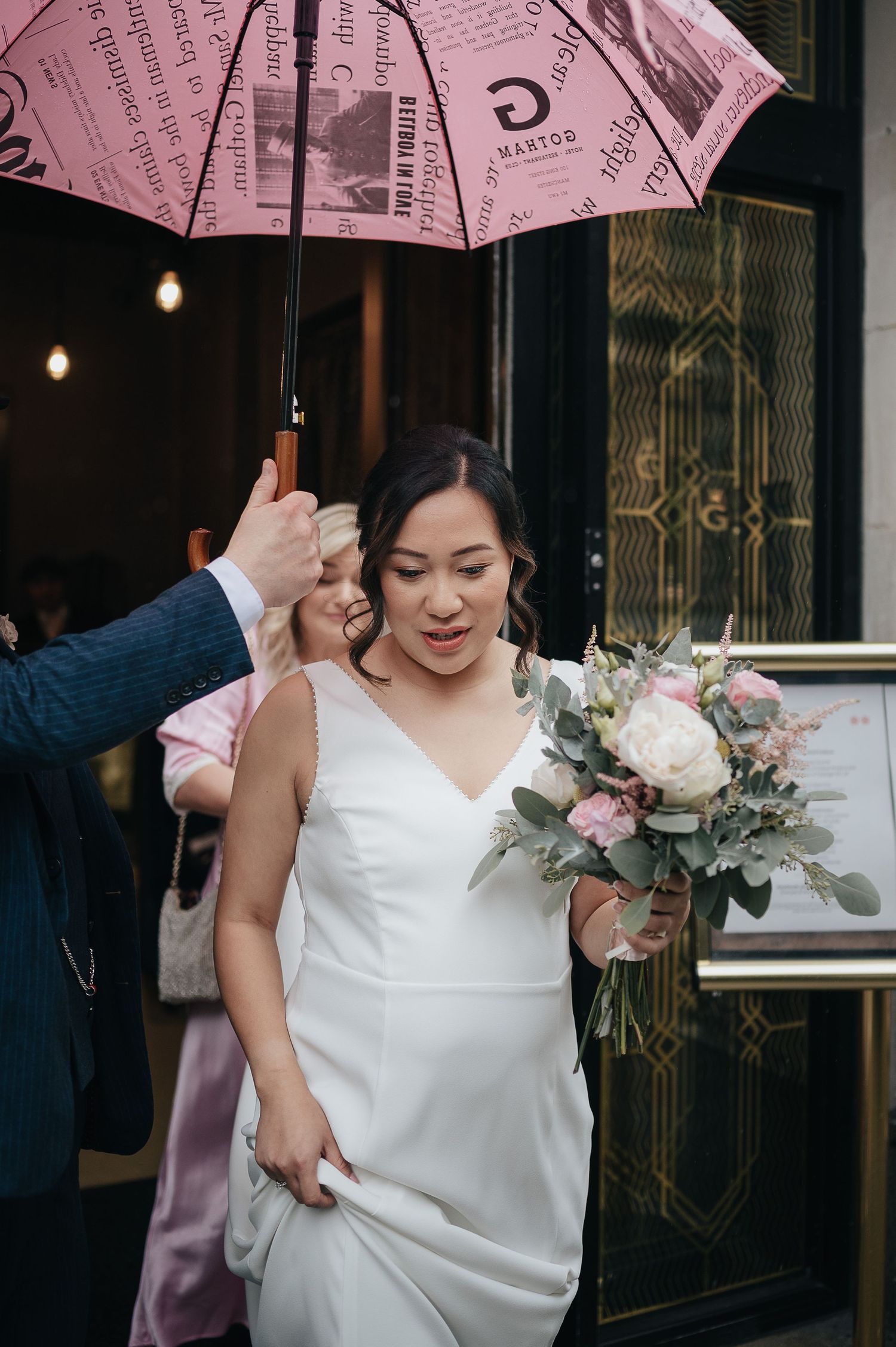Pink umbrella sheltering someone in a white wedding dress holding a floral bouquet.