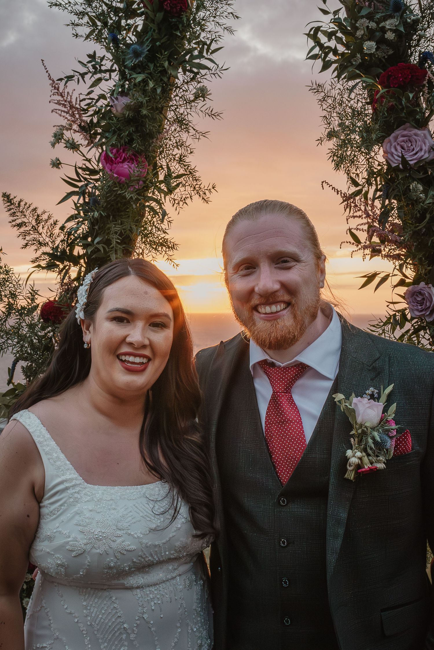 Wedding couple smiles together at sunset under a floral arch with purple and red flowers.