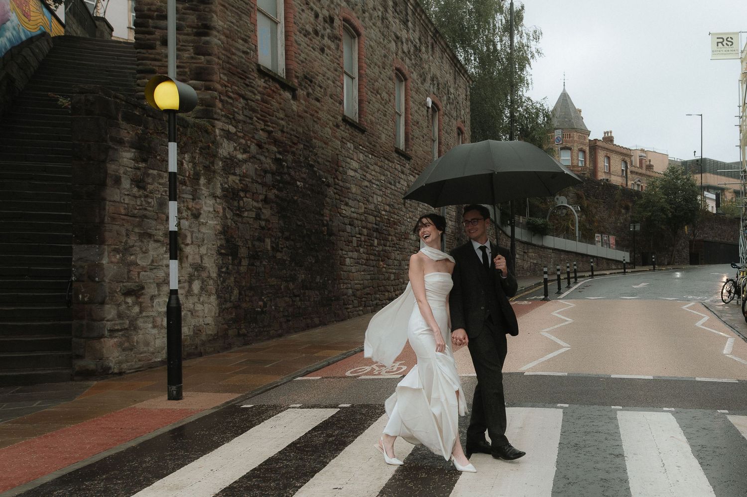 A couple walks along a historic cobblestone street with stone walls and lamp posts on a cloudy day.