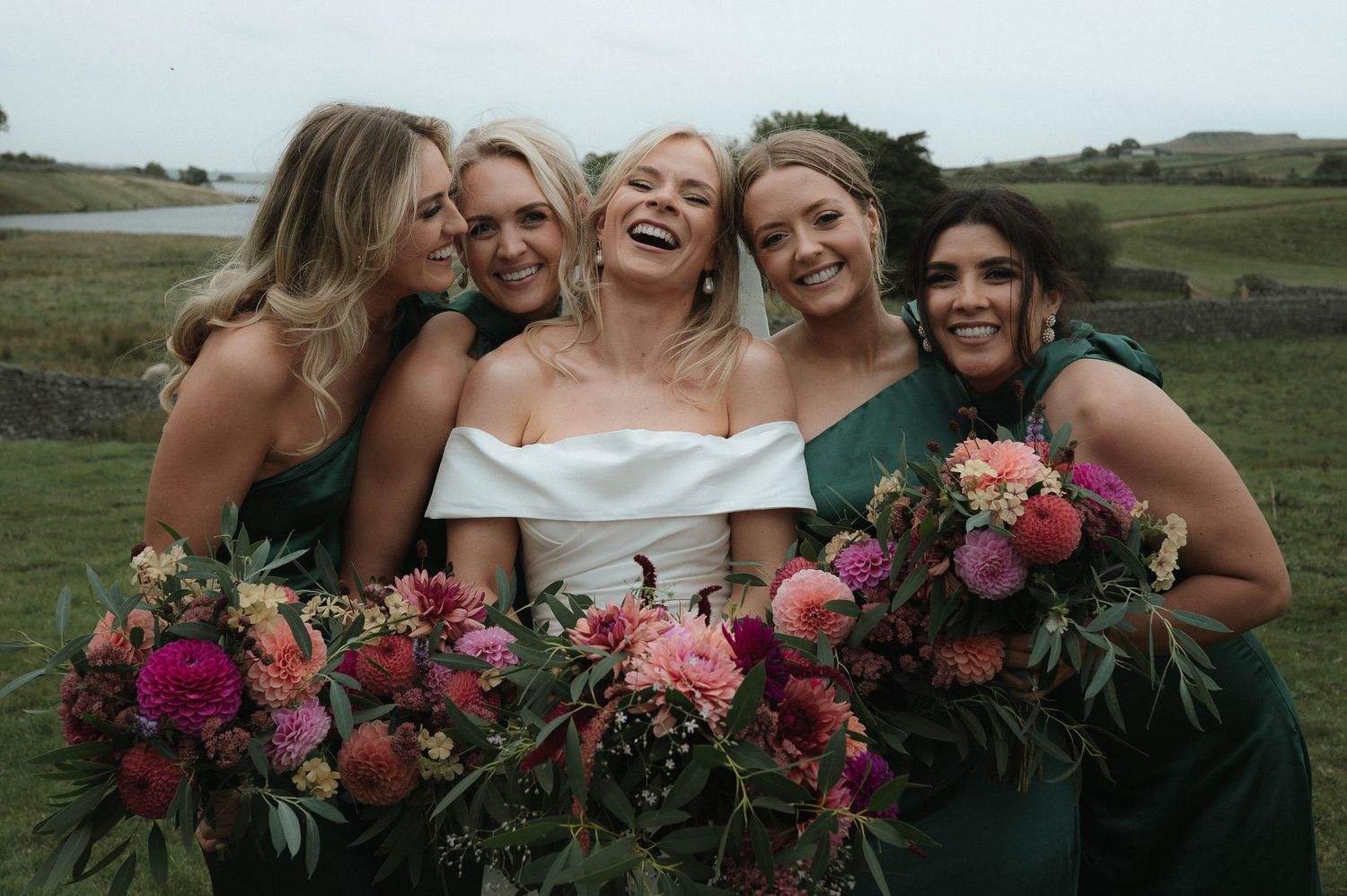 Wedding party holding vibrant pink and burgundy floral bouquets against scenic landscape. Blackton Grange Wedding. Luke Hassall Photography.