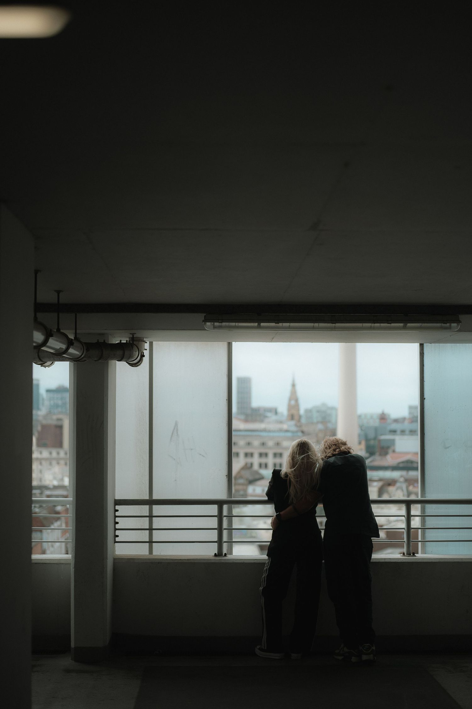 Two silhouettes stand in a dimly lit parking garage overlooking a cityscape through large windows.