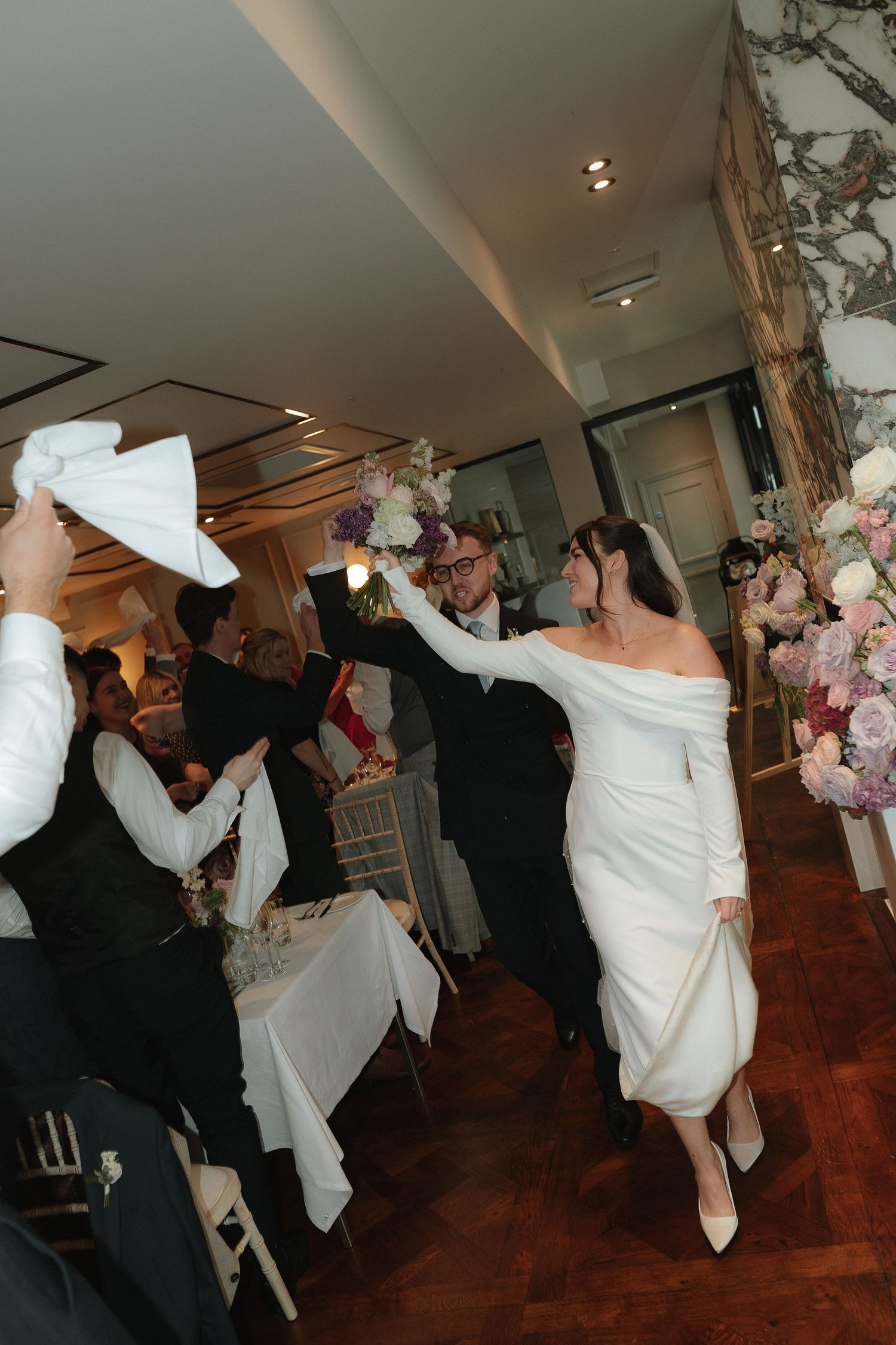 Bride and groom make grand entrance at elegant wedding reception, guests waving white napkins in celebration.