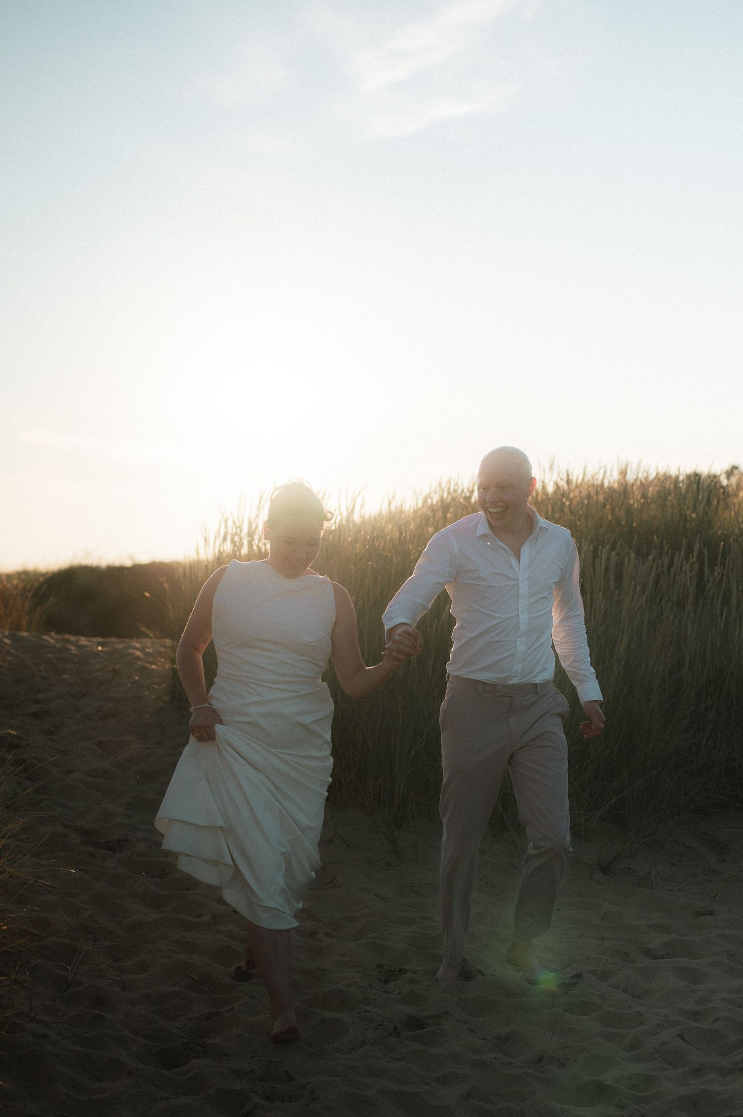 A couple walks together on a beach path at sunset with tall grass silhouetted against the golden sky.