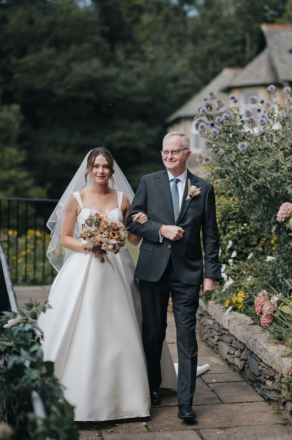 A classic wedding moment as the bride and companion walk down a stone path lined with flowers and greenery.