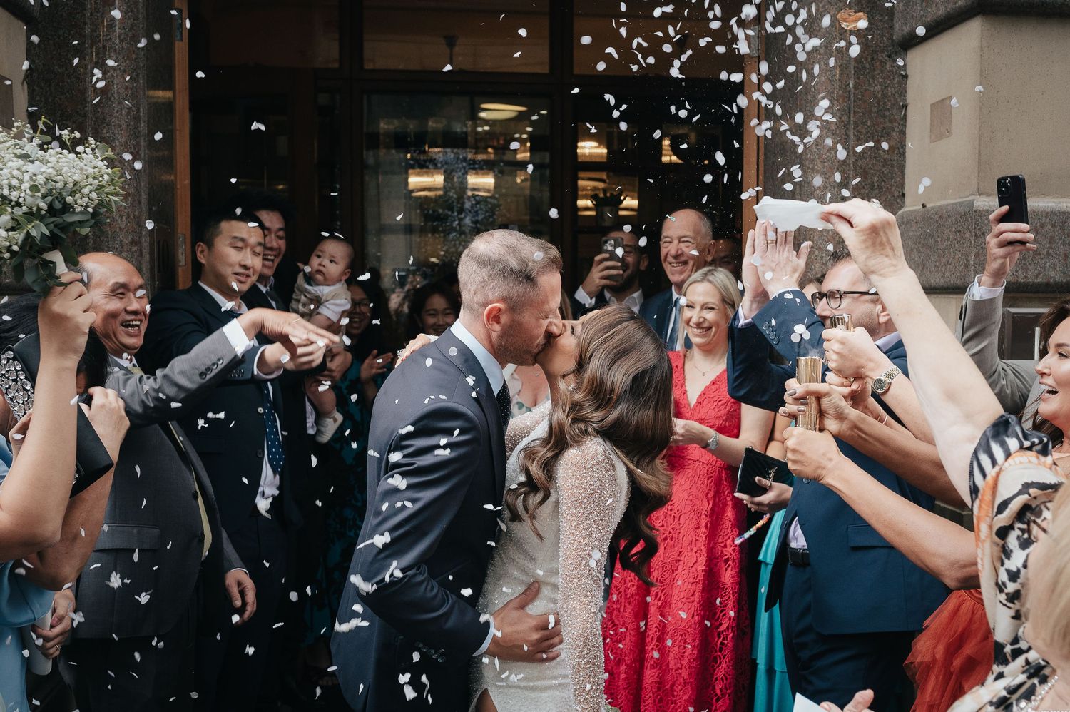 Wedding party exits venue as guests throw confetti in celebration.