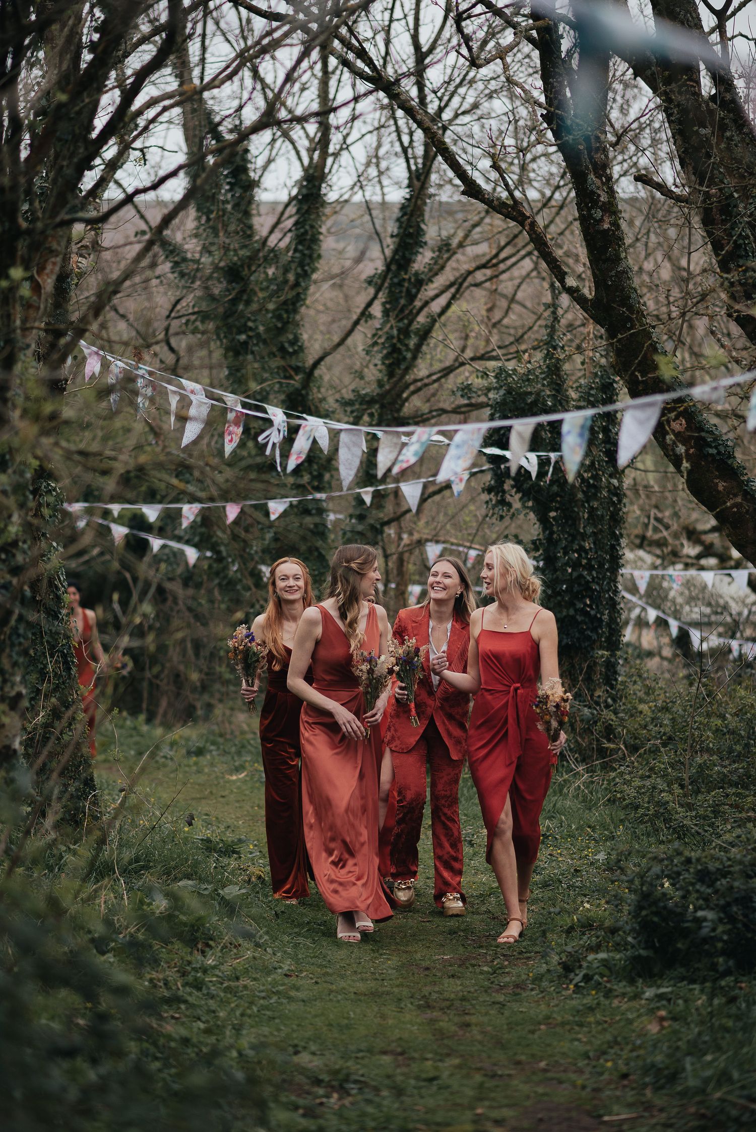 Bridesmaids in red dresses walk along a garden path decorated with white bunting.