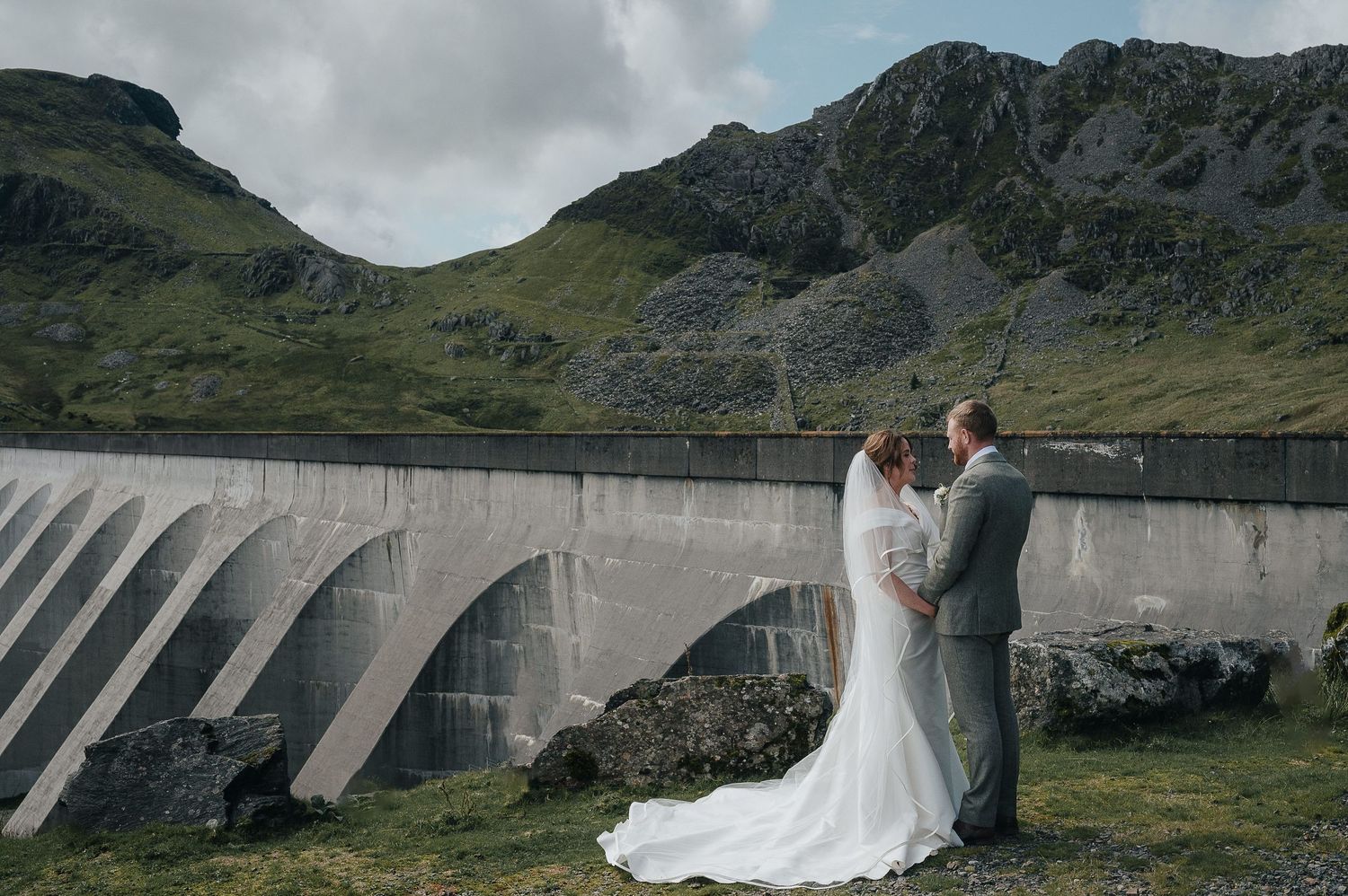 A couple in wedding attire poses romantically in front of a large concrete dam surrounded by scenic mountain landscape.