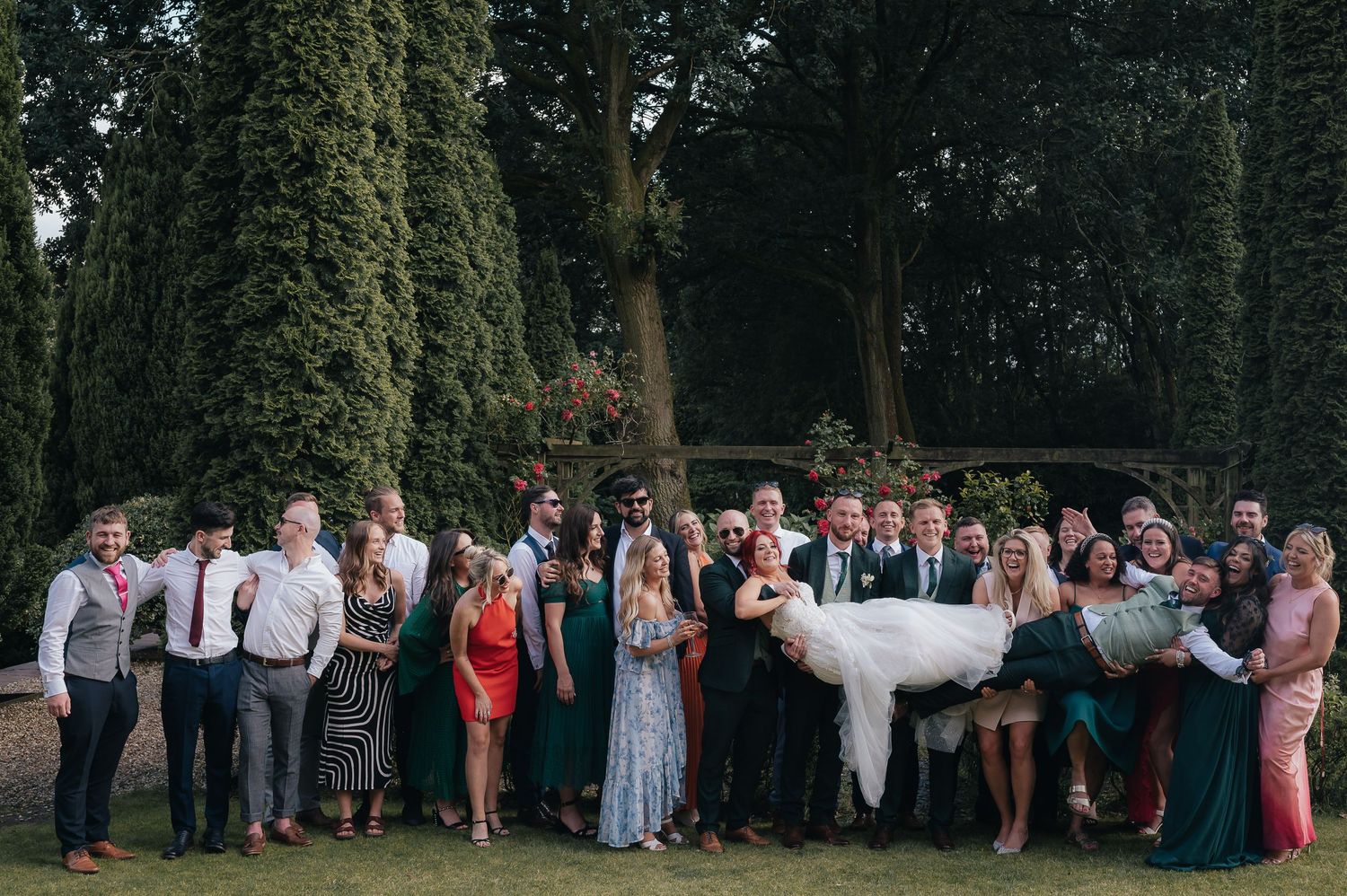 A large wedding party poses together outdoors in formal attire with greenery and trees in the background.