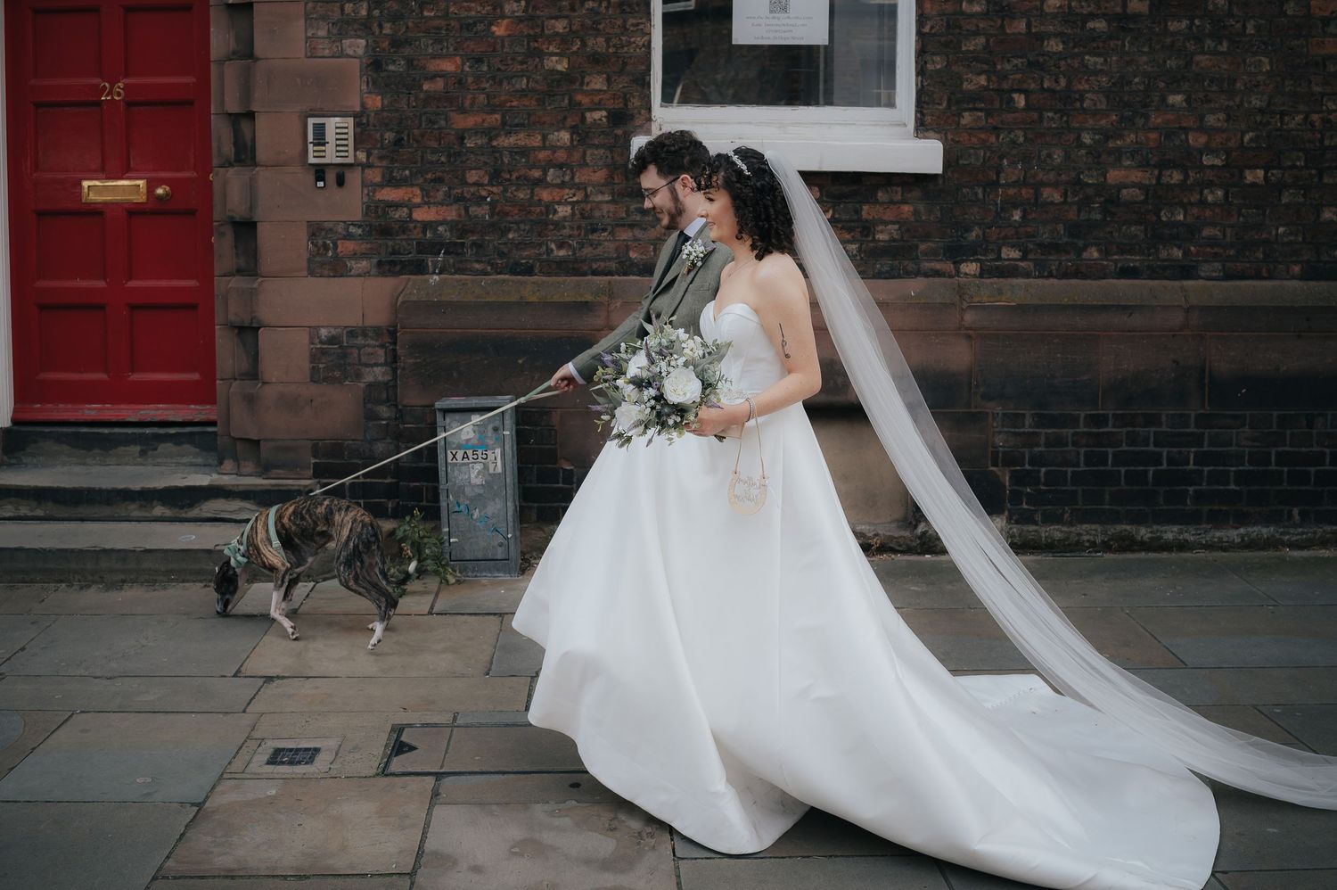 A bride in a flowing white dress with a dog on a leash near a red door.
