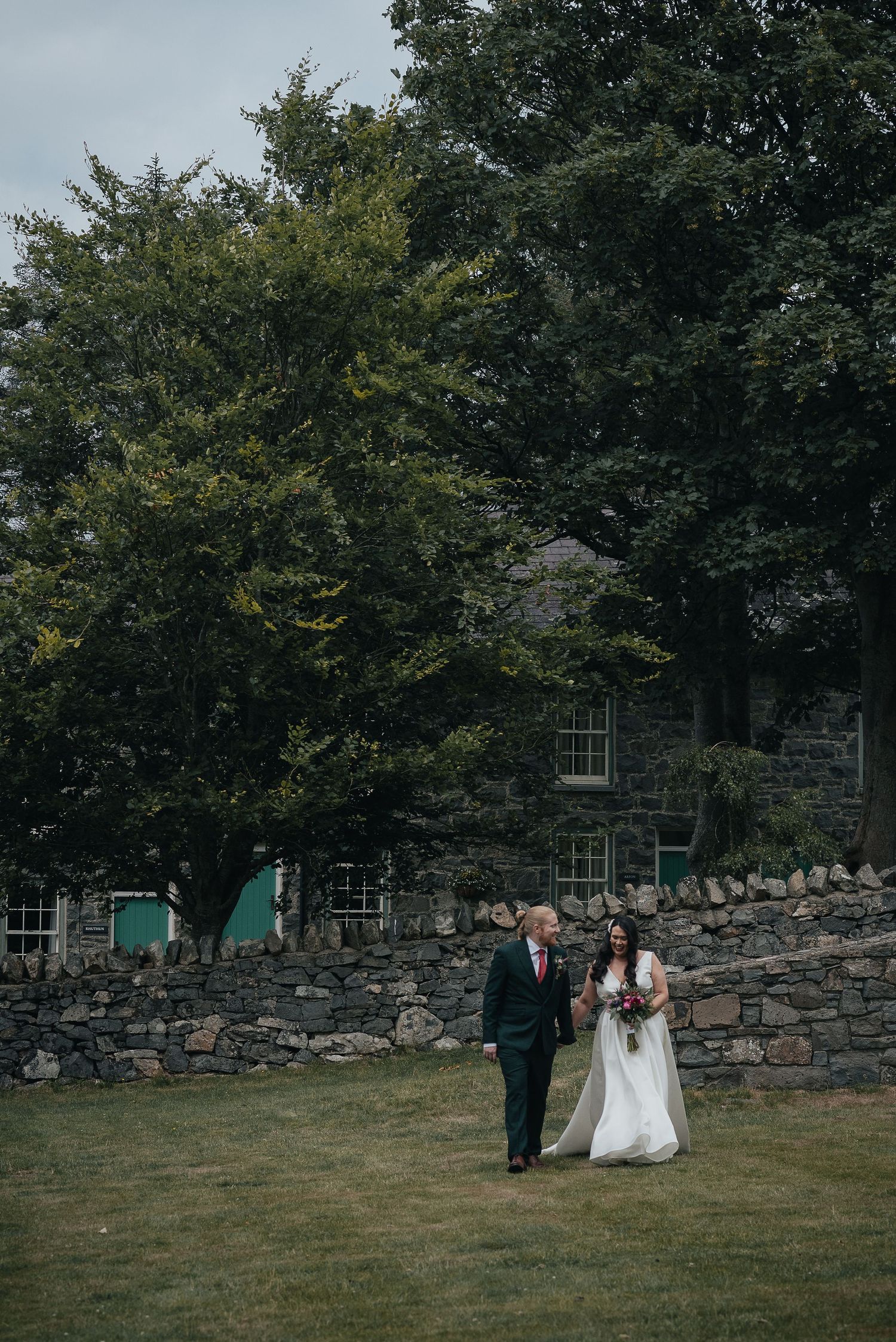 A couple walking on grass against a rustic stone building backdrop with lush green trees.