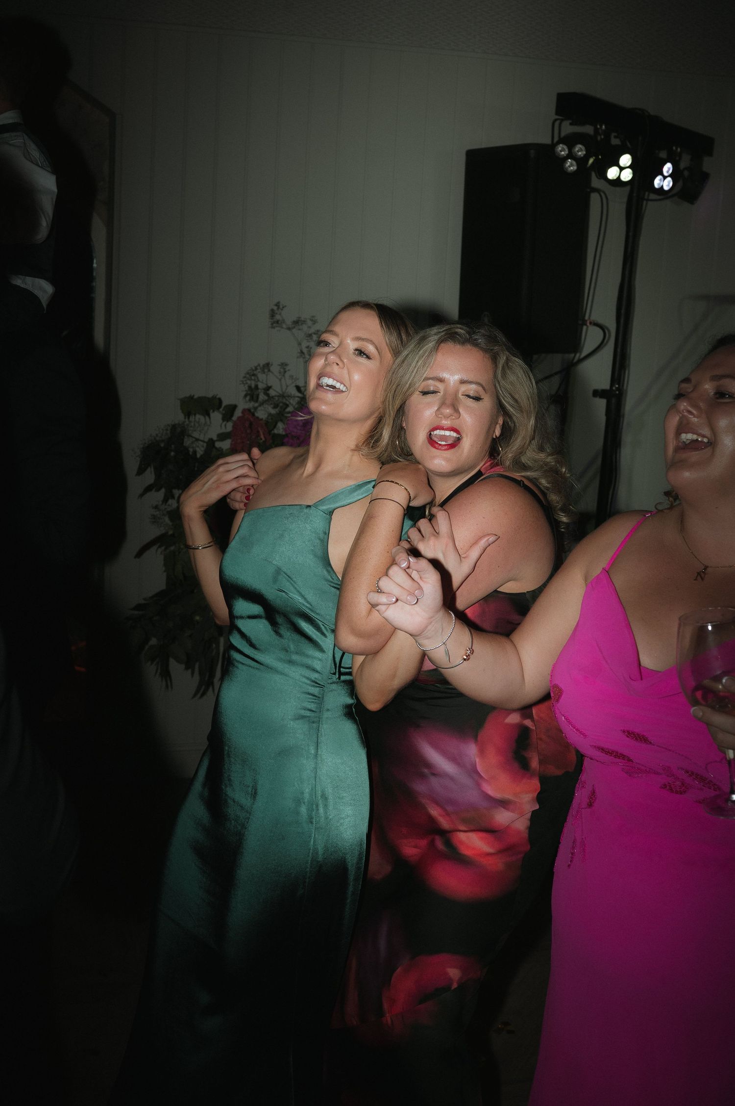 Guests in bright dresses dancing and laughing at a dimly lit wedding reception.