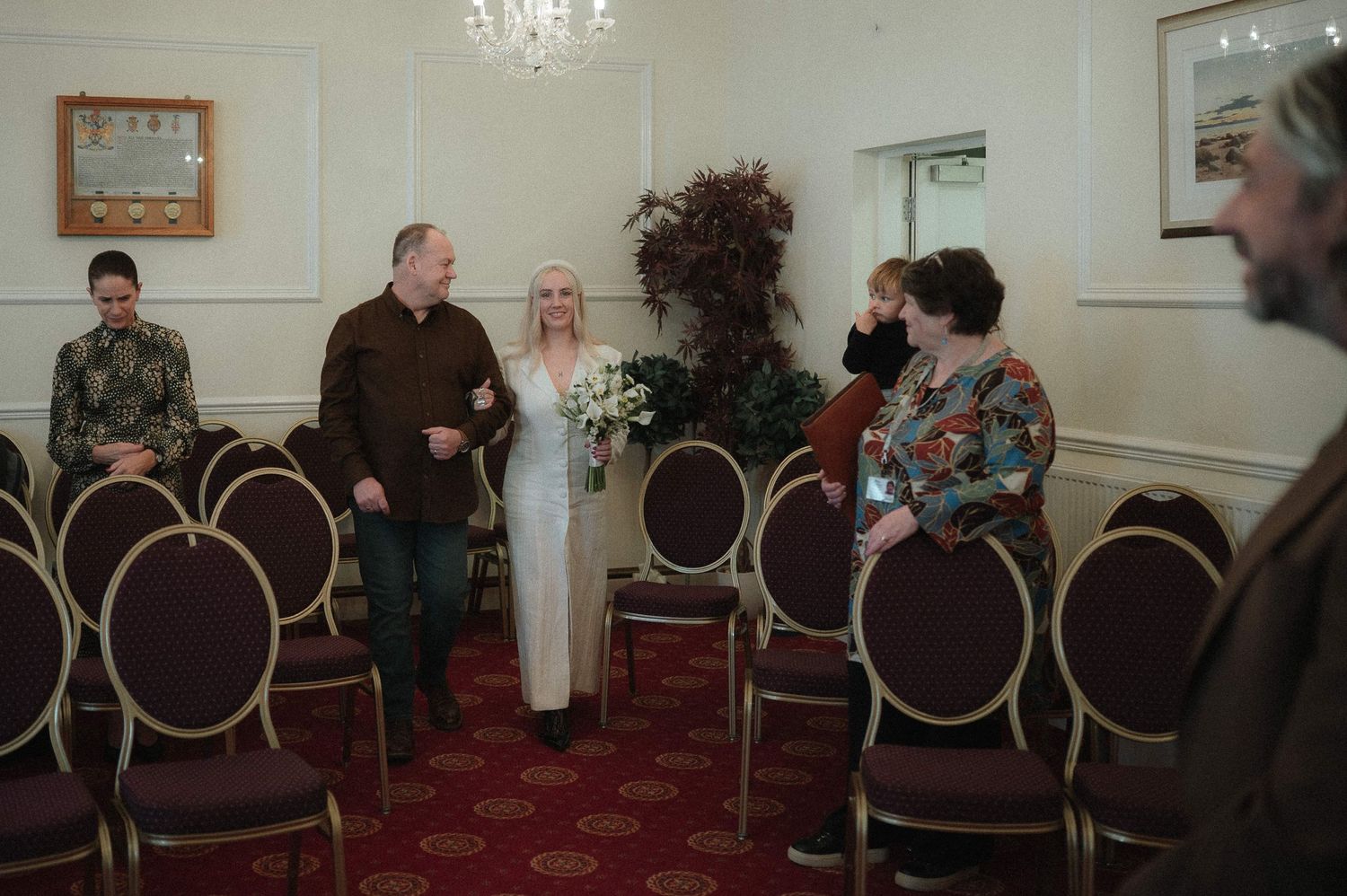 A small wedding ceremony takes place in an intimate room with round-backed chairs and white walls.