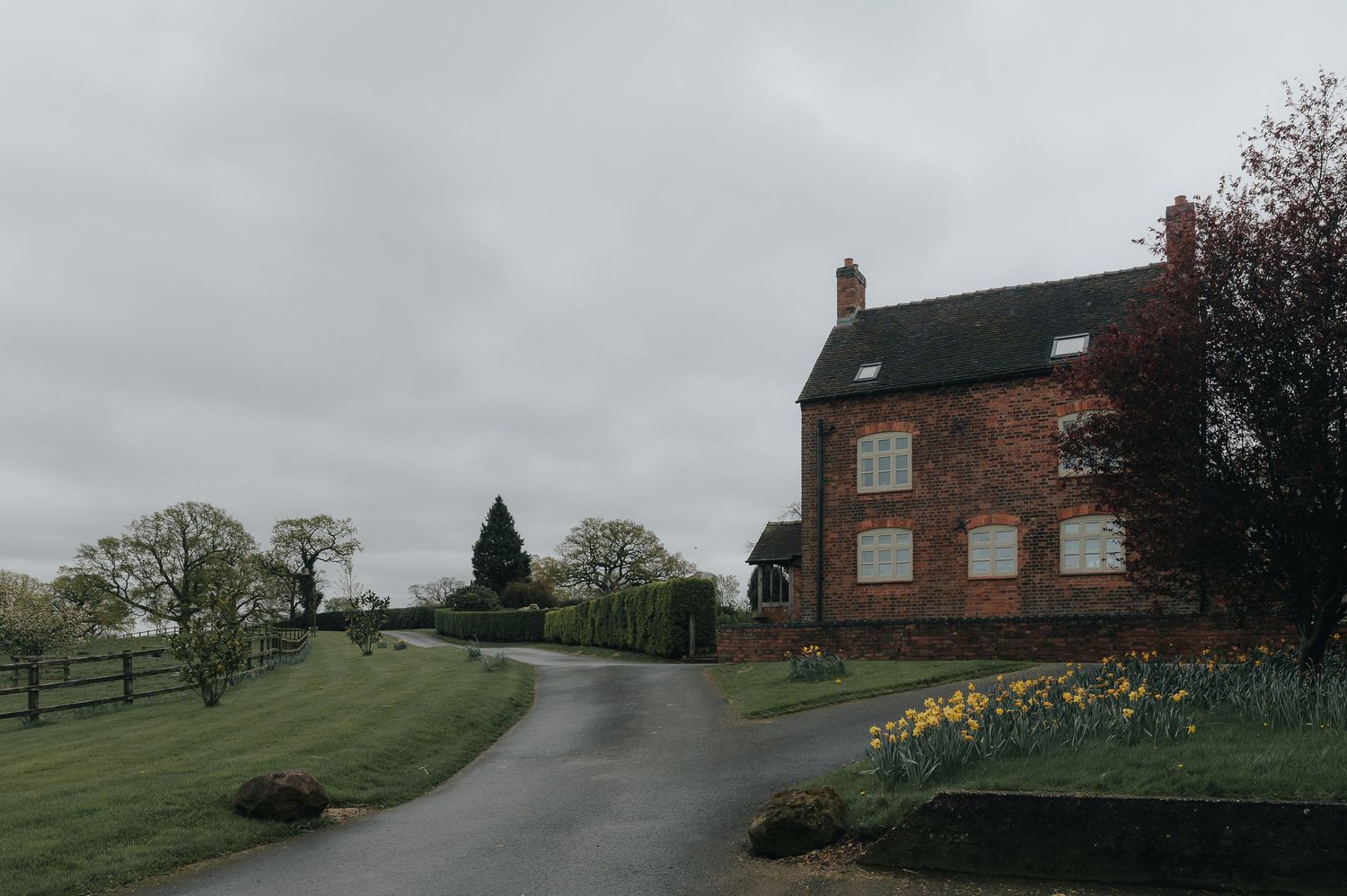 Red brick farmhouse on a green countryside hill with daffodils and winding path.