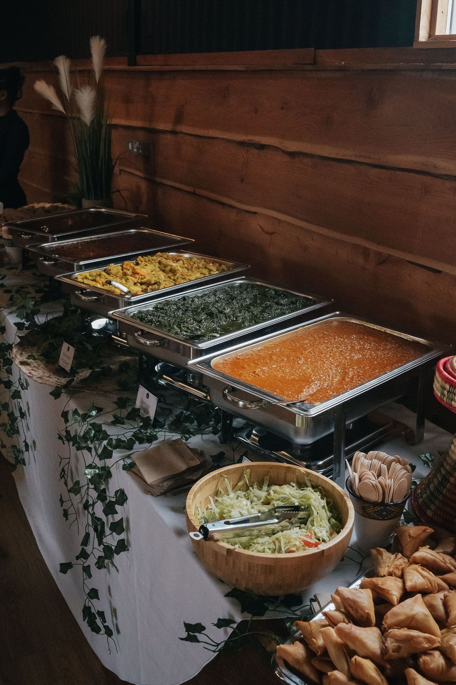 Wedding buffet spread with colorful dishes in chafing dishes against wooden wall.