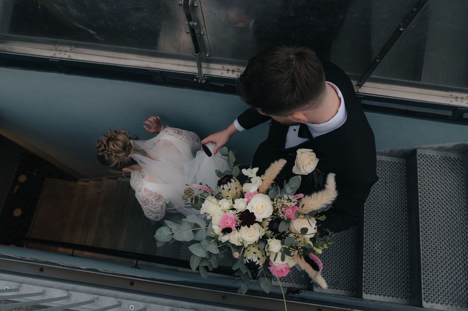 Overhead view of someone in formal attire holding a blush pink and white floral bouquet on modern grey stairs.