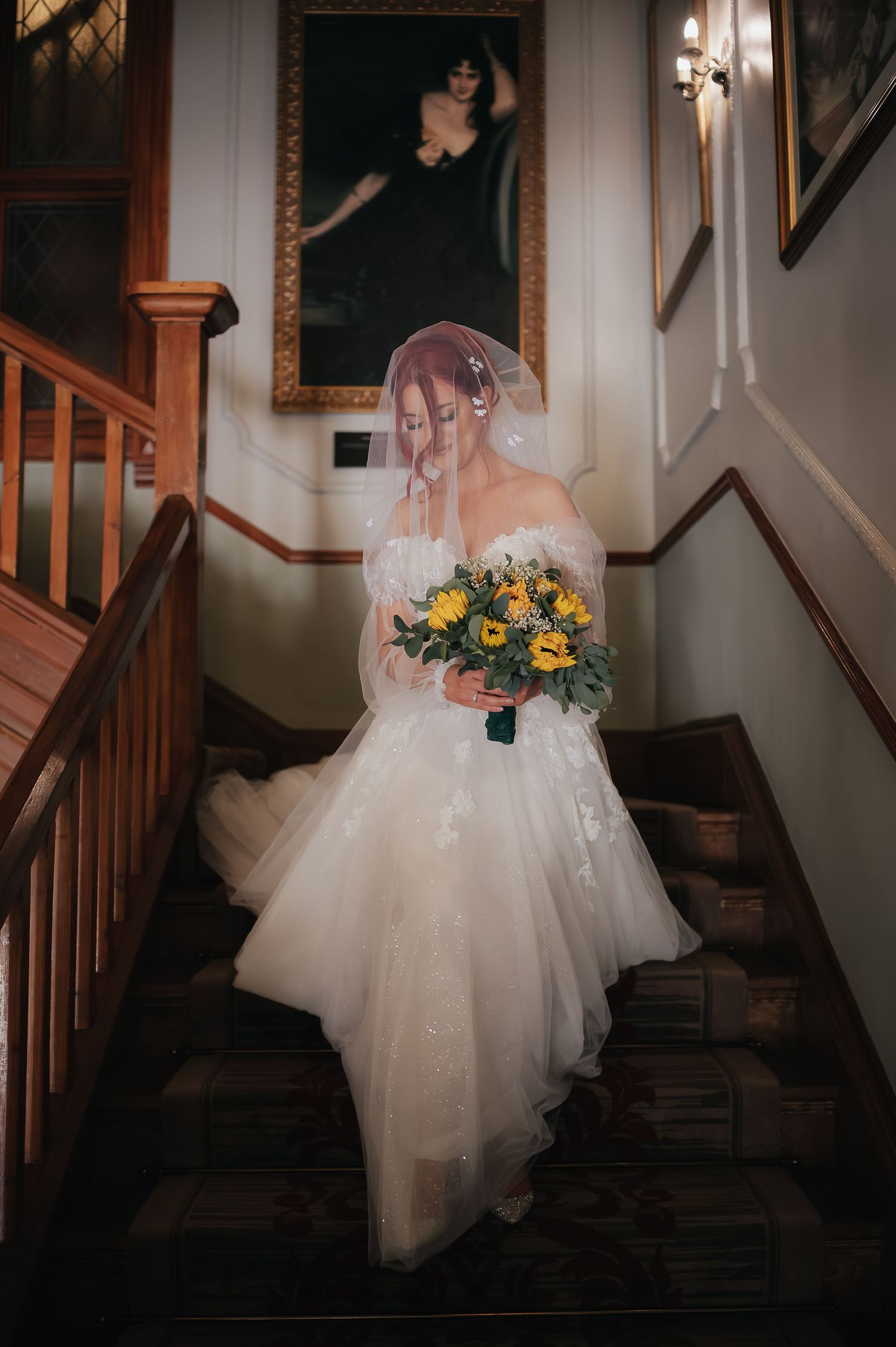 Bride in flowing white gown poses on wooden staircase while holding sunflower bouquet.