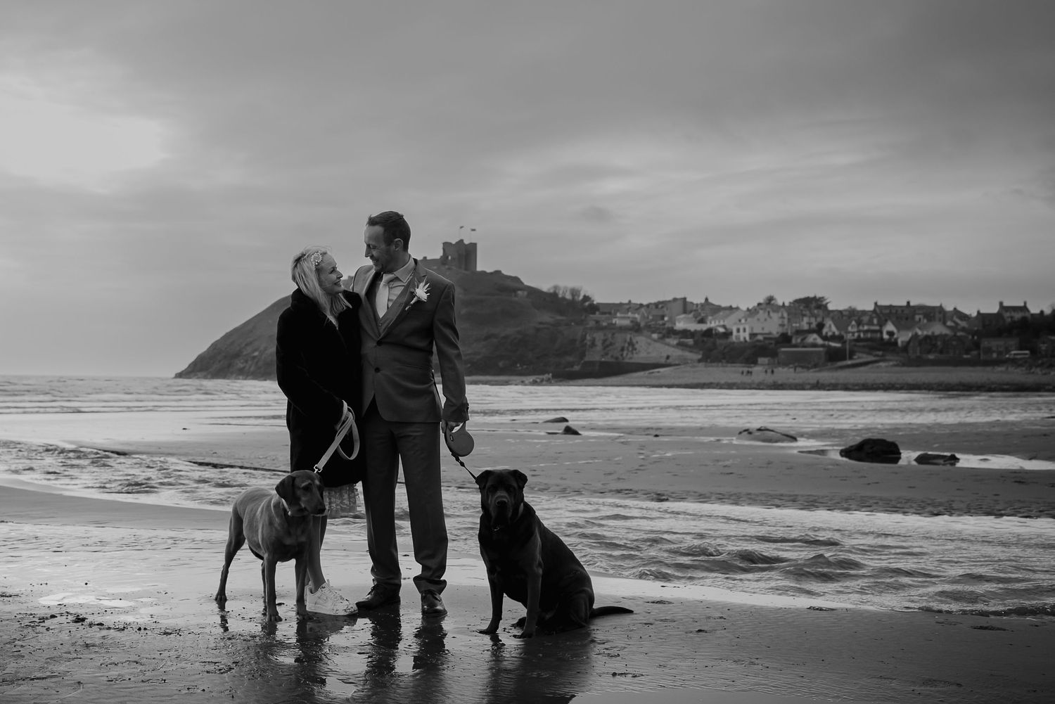 A couple stands on a beach with two dogs against a dramatic coastal landscape with a castle in the background.