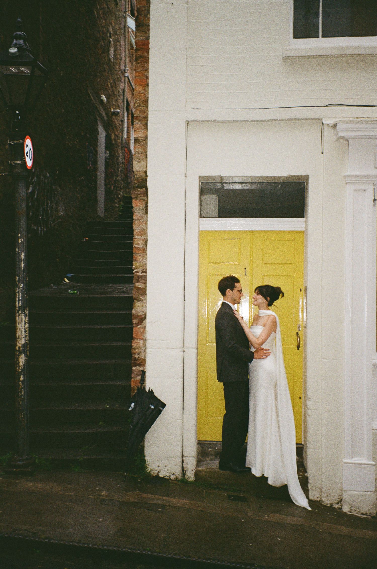 Bride and groom embrace before a bright yellow door on a charming urban street in a romantic wedding portrait.