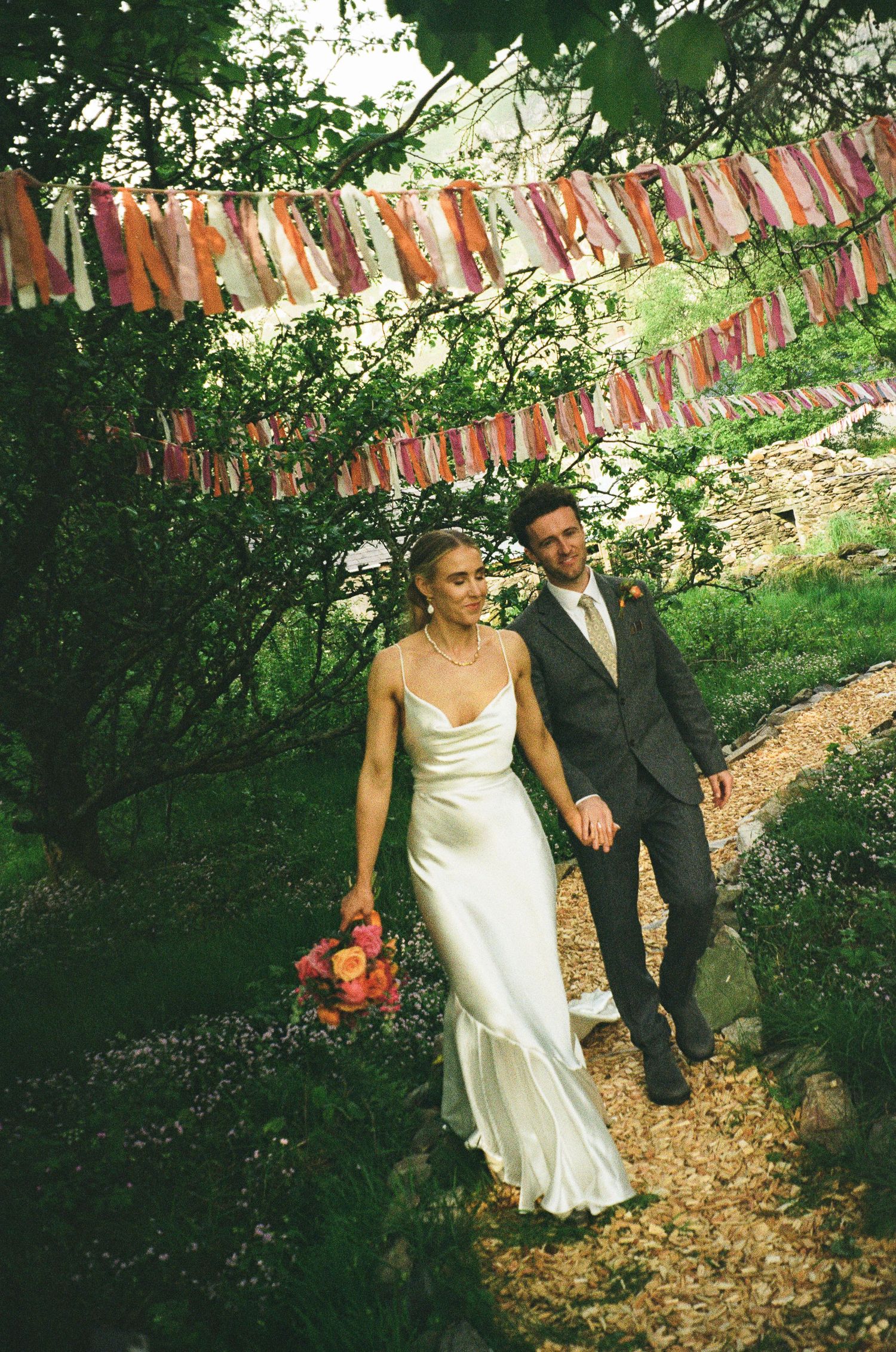 Bride and groom walk hand-in-hand through a garden path adorned with colorful ribbon bunting at their outdoor wedding.