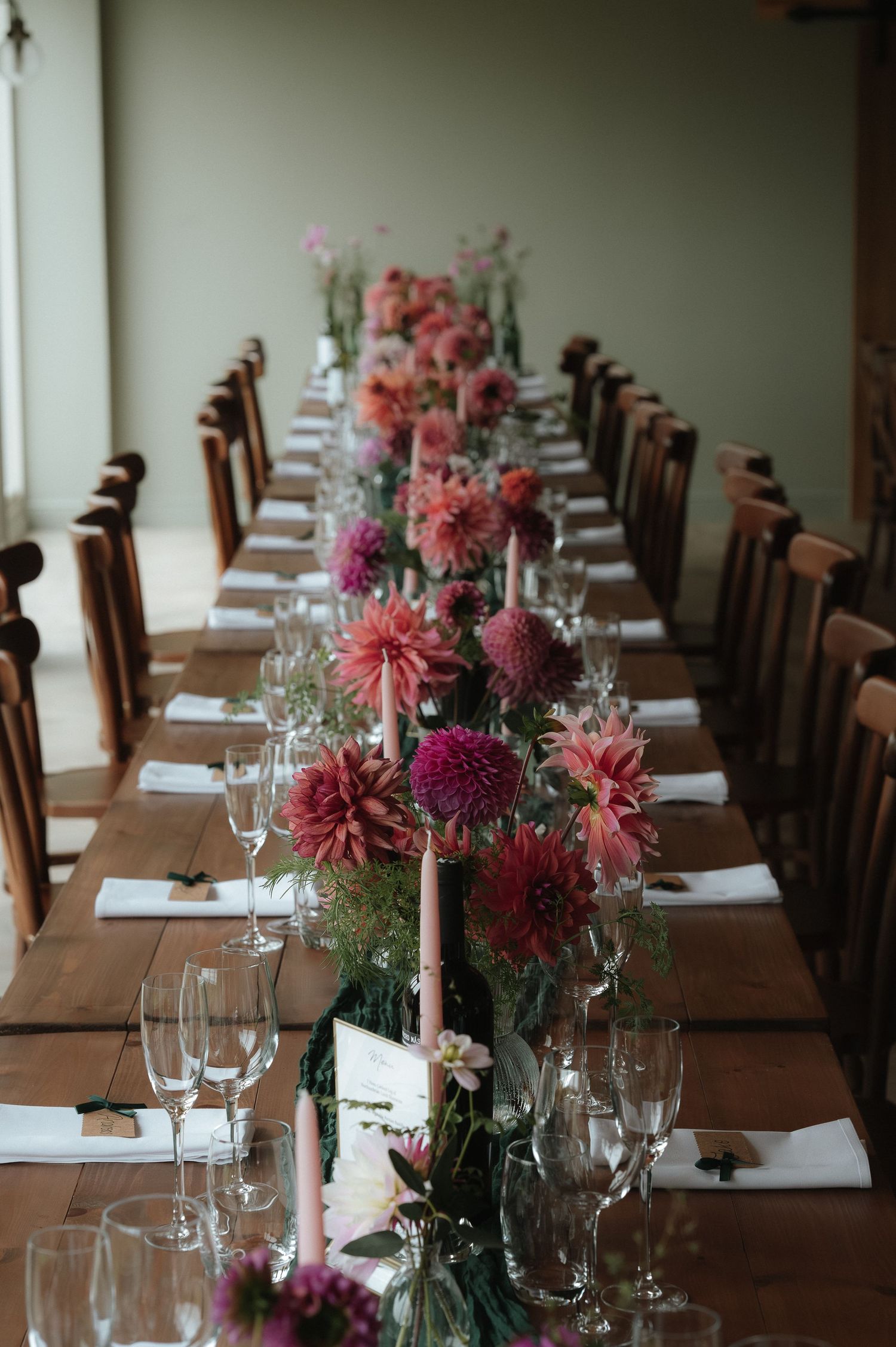 Elegant wedding table setting with pink and burgundy dahlia floral centerpieces and wooden chairs at a rustic reception.
