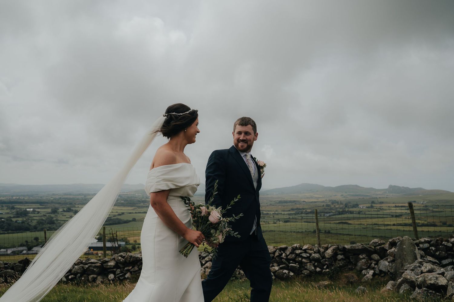A wedding couple stands against a dramatic cloudy landscape with mountains in the background.
