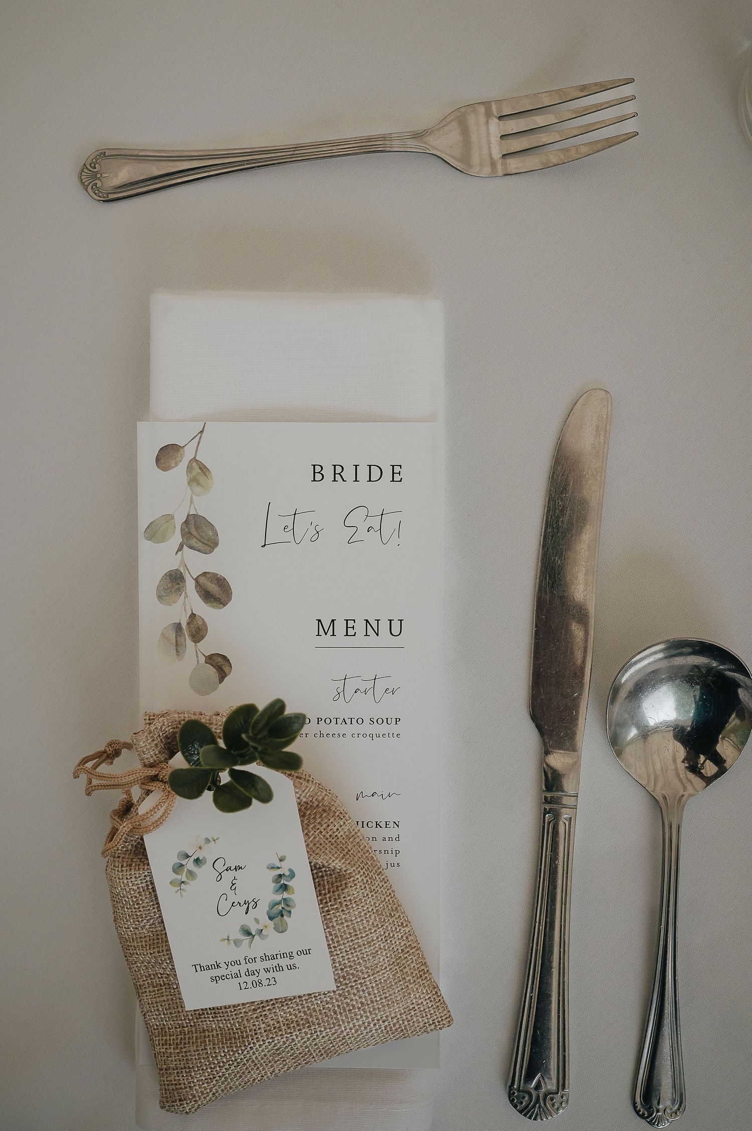 Wedding reception place setting with menu card, silverware, and rustic burlap favor bag.