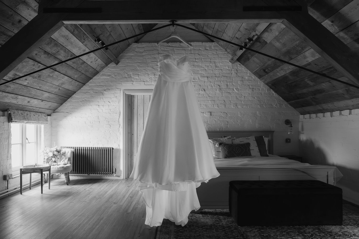 Wedding dress hanging in attic room with white brick walls and wooden beams.